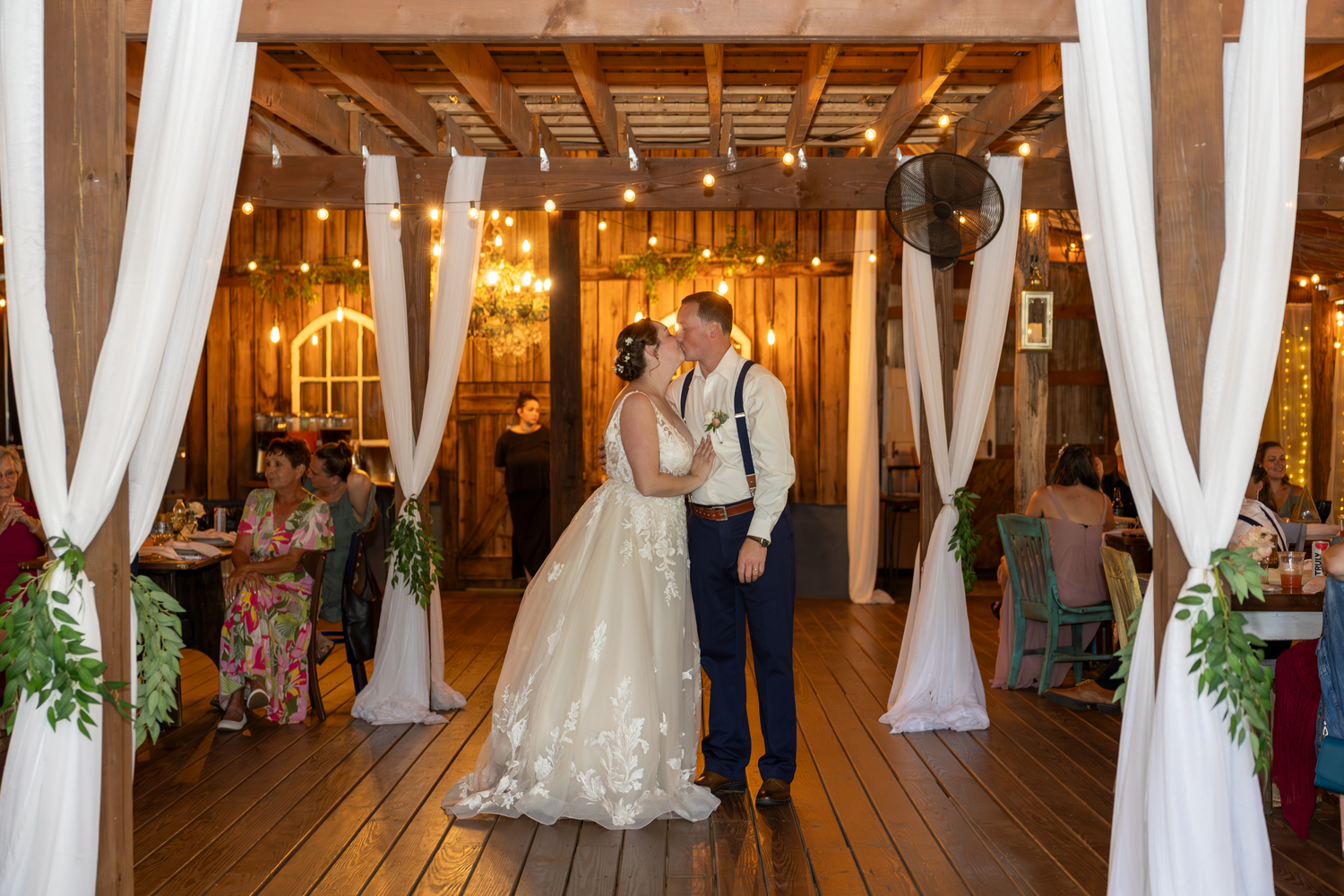 Bride and groom share first dance in rustic barn wedding venue with white drapes and string lights.