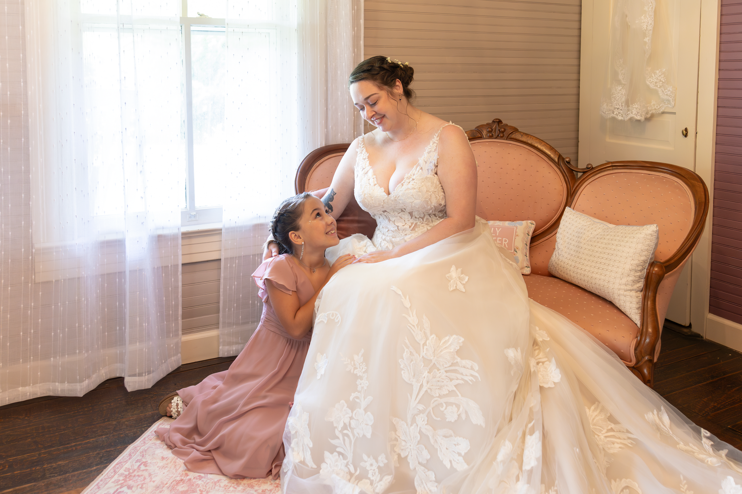 Bride in lace wedding gown sits on vintage sofa as flower girl in pink dress looks up at her adoringly.