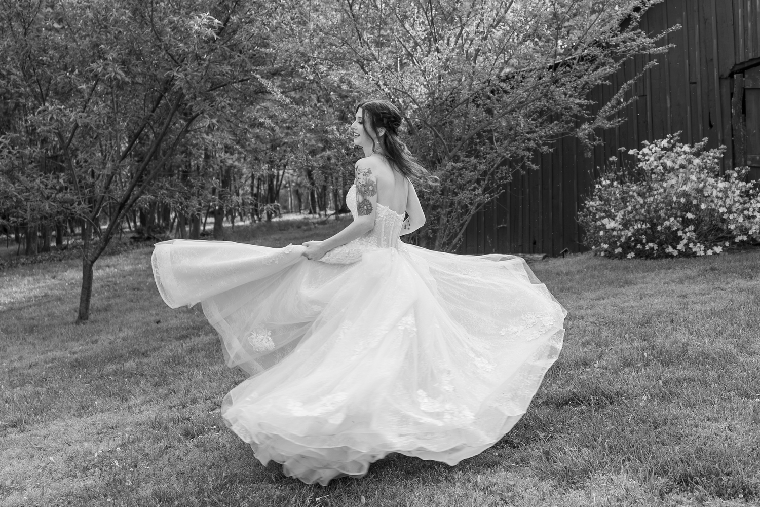 Bride twirling in flowing white wedding gown outdoors in garden setting, black and white photography.