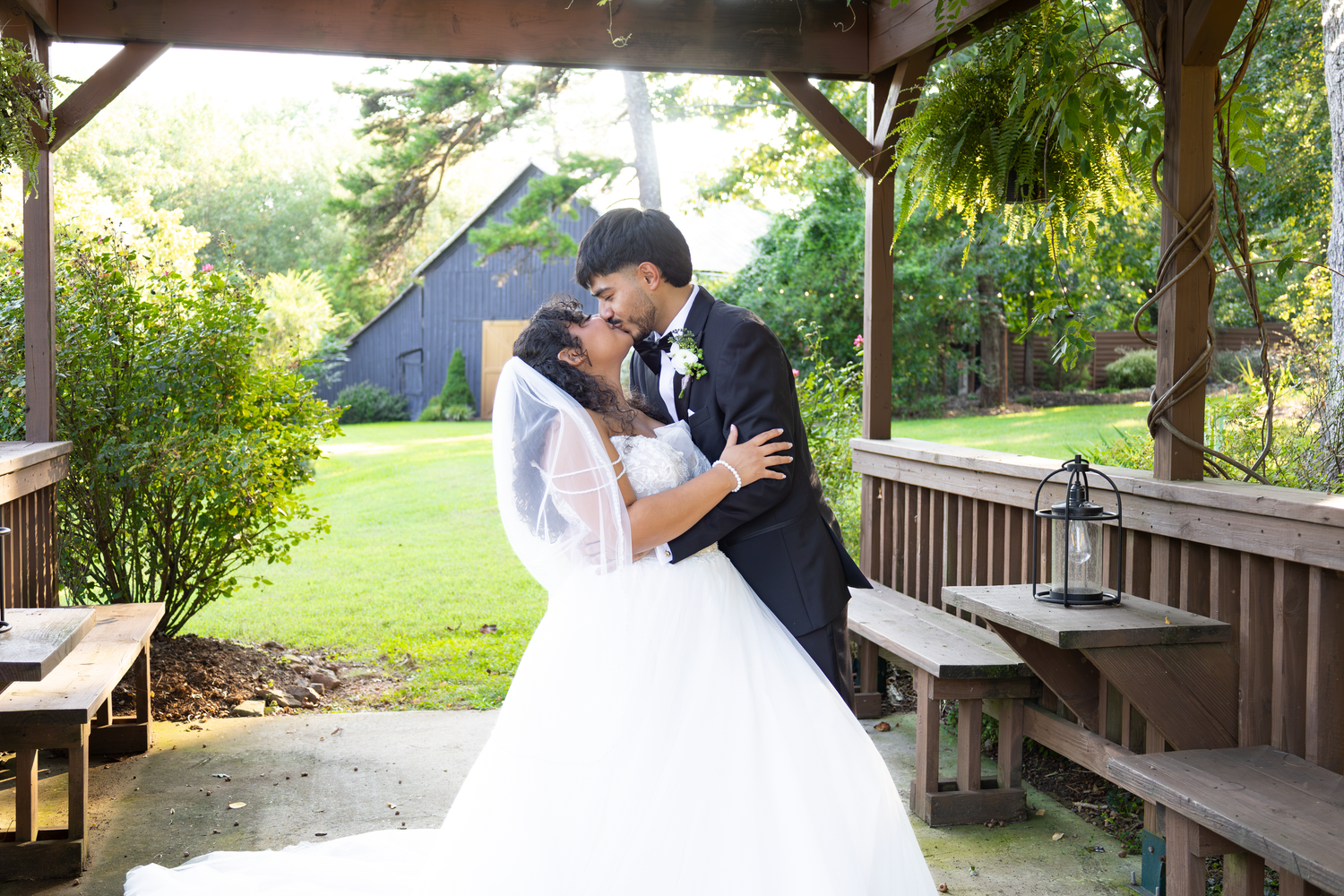 Bride and groom share a romantic kiss under a wooden gazebo surrounded by lush greenery on their wedding day.