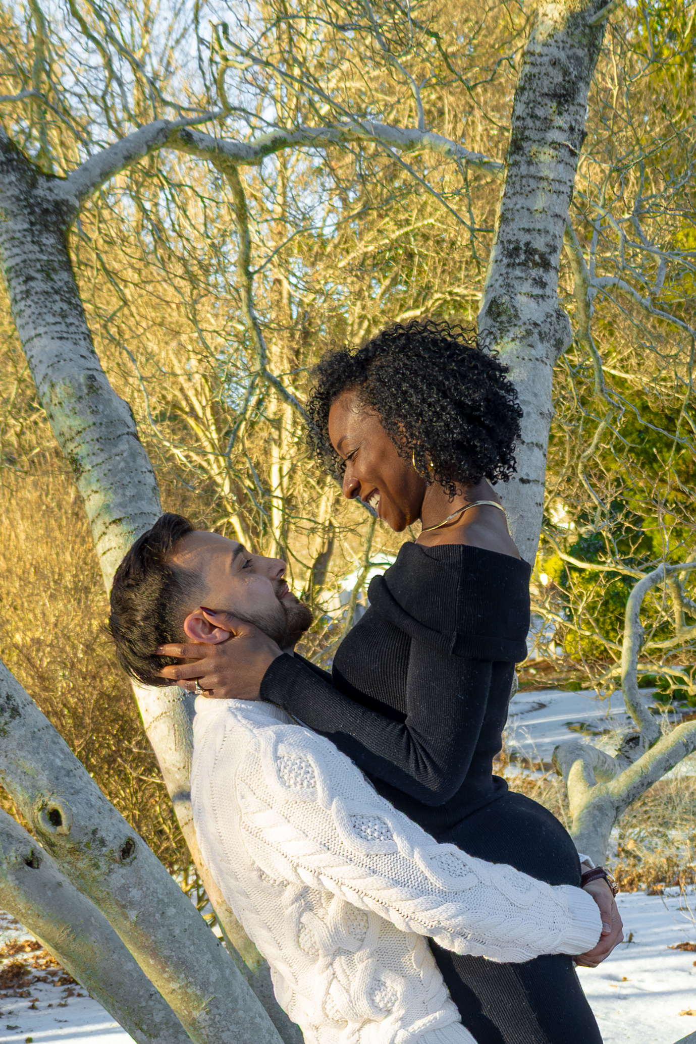 A couple shares a romantic moment near a birch tree in winter sunlight.