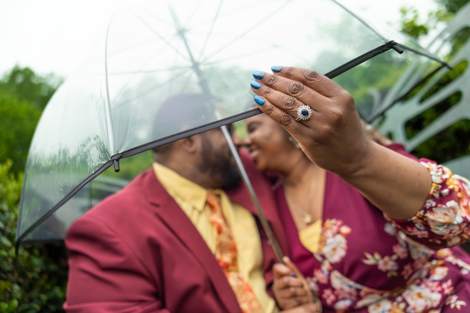 Couple sharing a clear umbrella in the rain, woman showcasing engagement ring with colorful nails, man in burgundy suit.