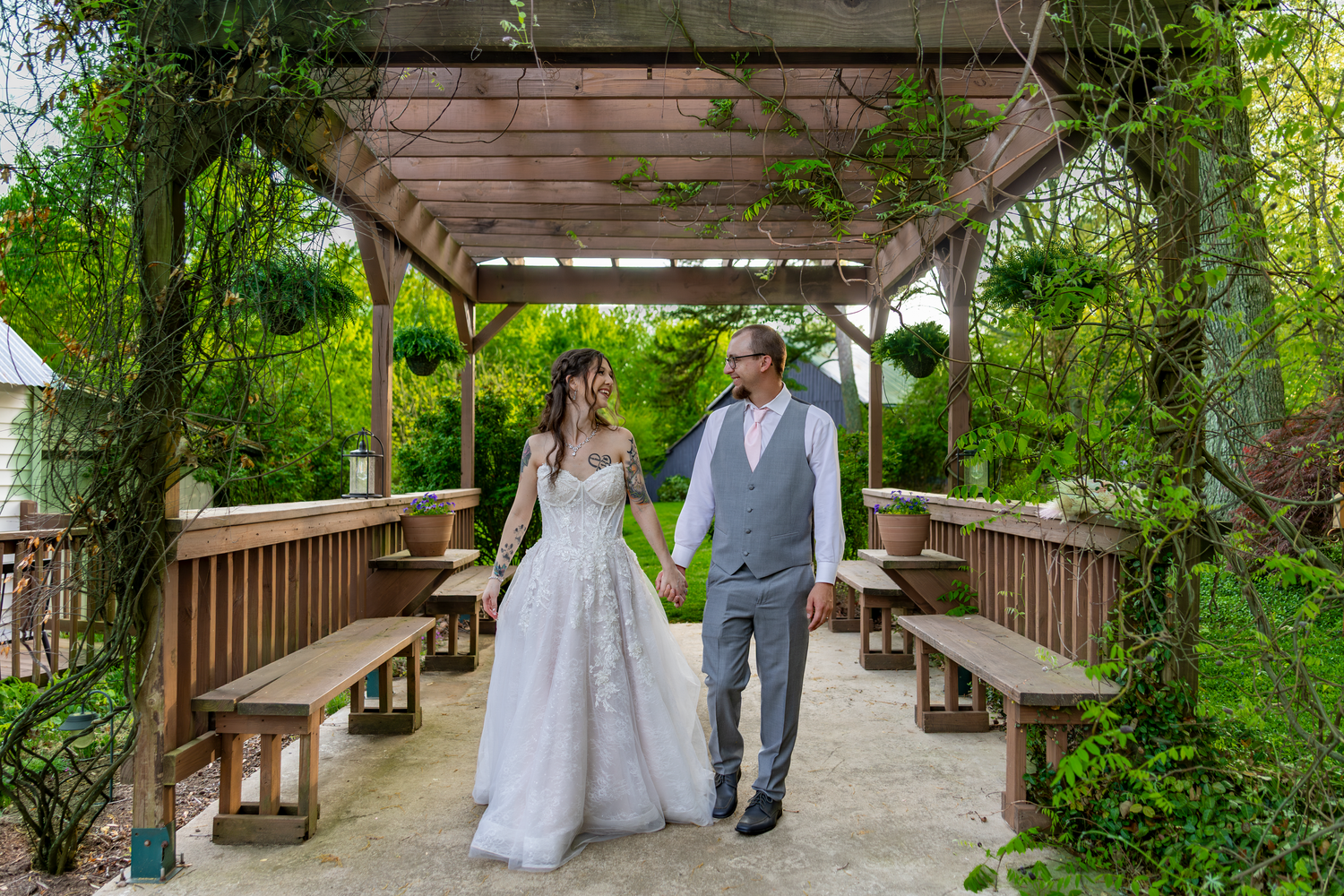 Bride and groom walk hand-in-hand under a wooden pergola surrounded by lush greenery at an outdoor wedding venue.