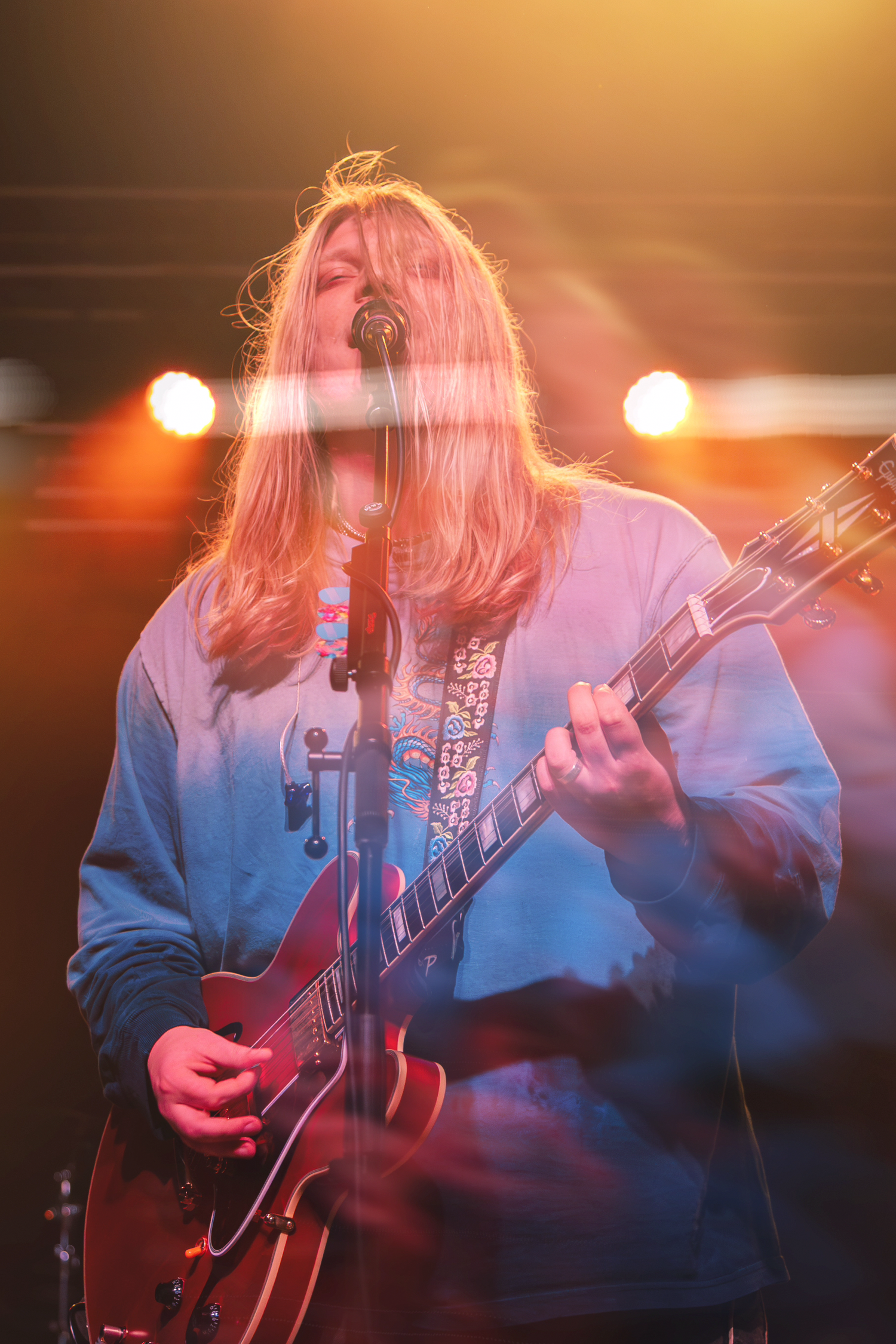 A musician performs on stage with a guitar under warm orange lighting during a live concert performance.