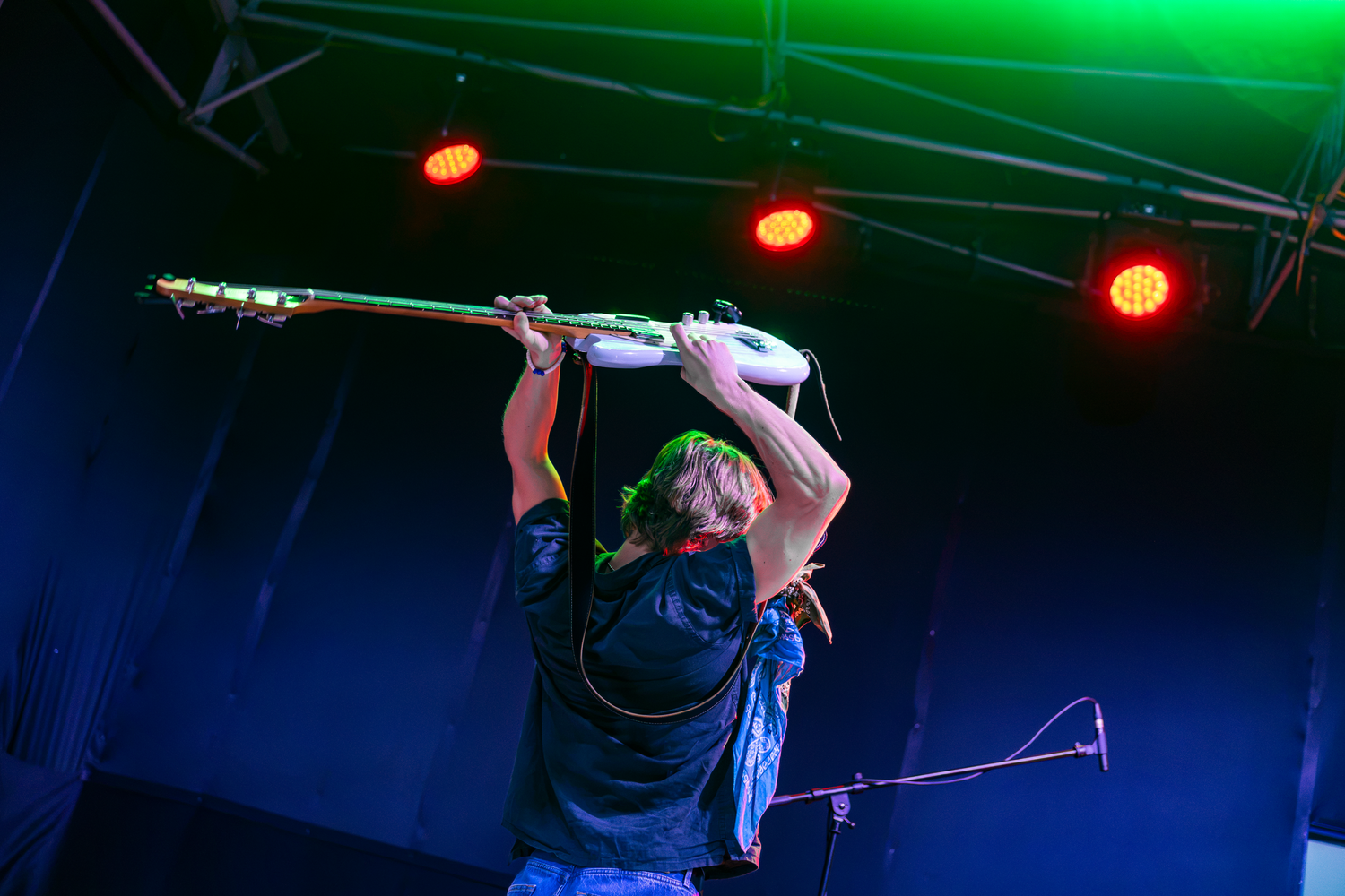 Guitarist raises electric guitar overhead on stage under red and green lights during live concert performance.