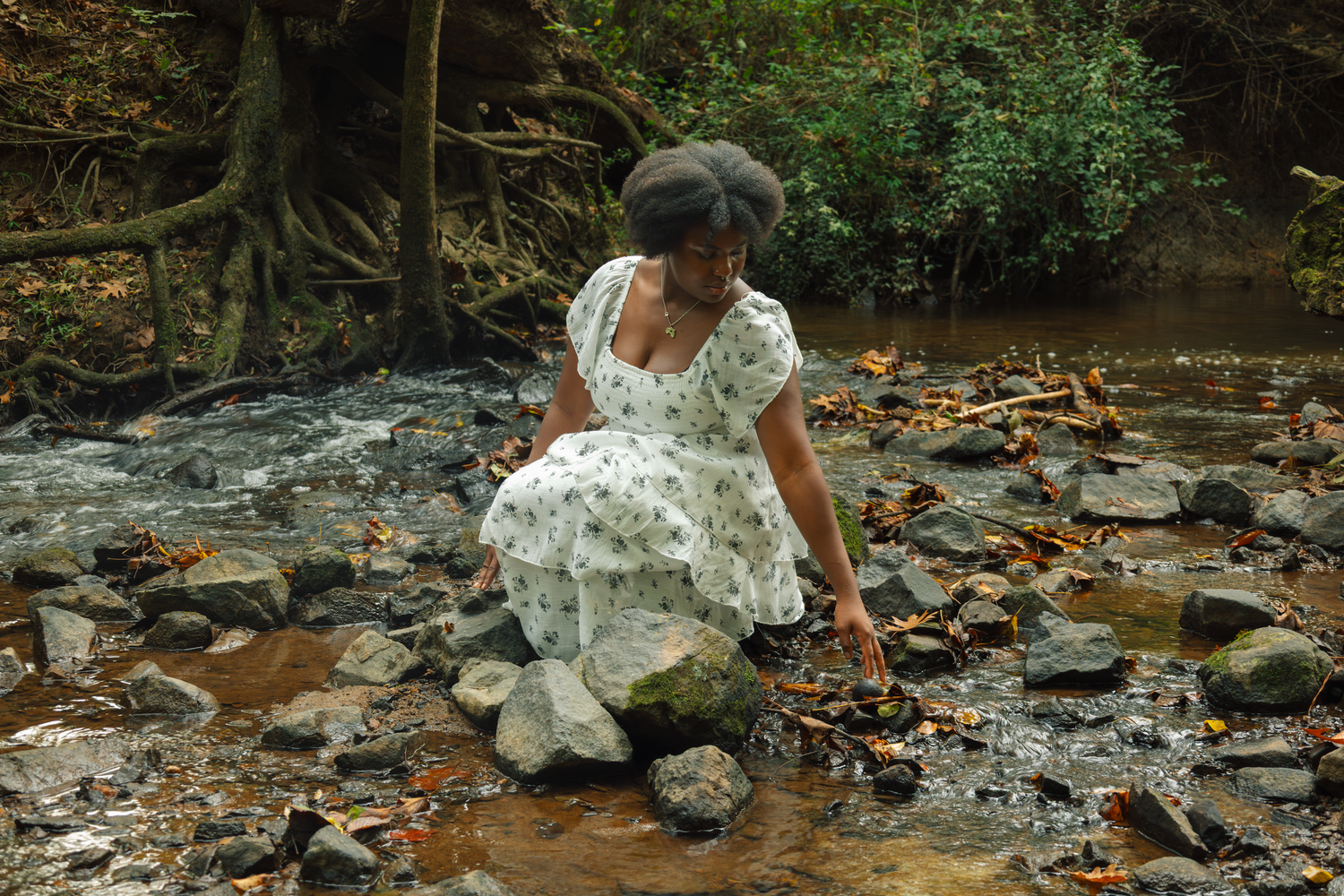 Woman in floral dress crouching by a rocky stream in a lush green forest, touching the water.