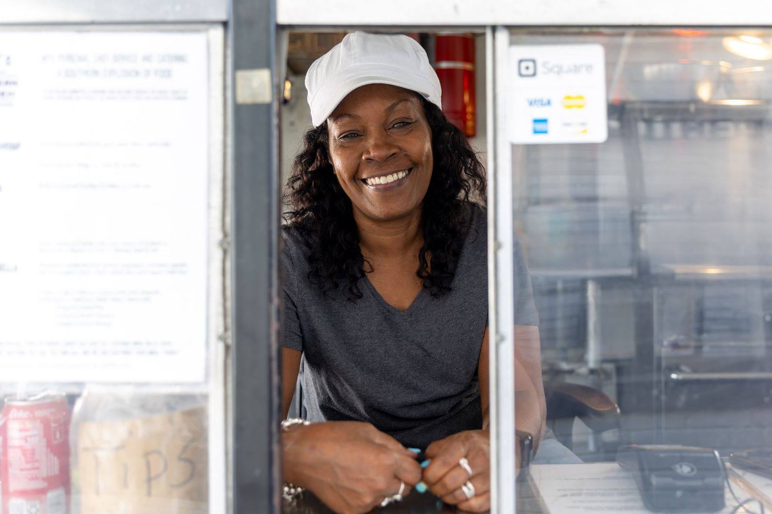 Smiling woman in white cap leaning out of food truck window with Square payment sign visible.