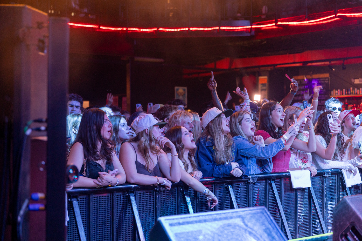 Concert crowd leans against a railing with red neon lights glowing above in a dark venue.
