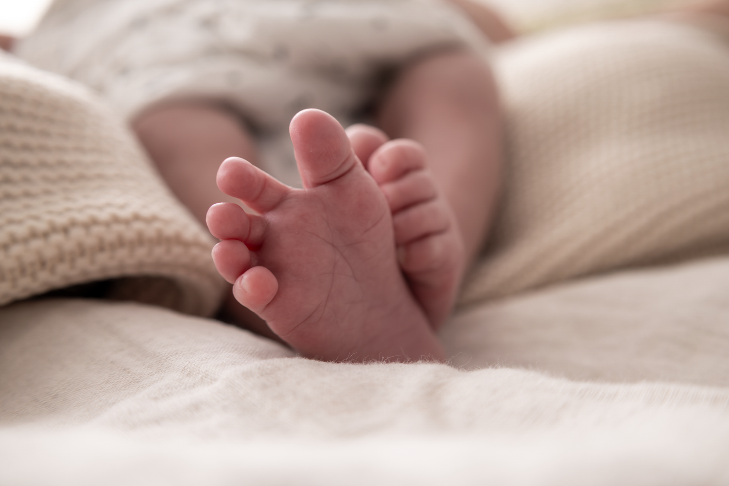Close-up photo of a newborn baby's tiny feet resting on soft white blankets in natural light.
