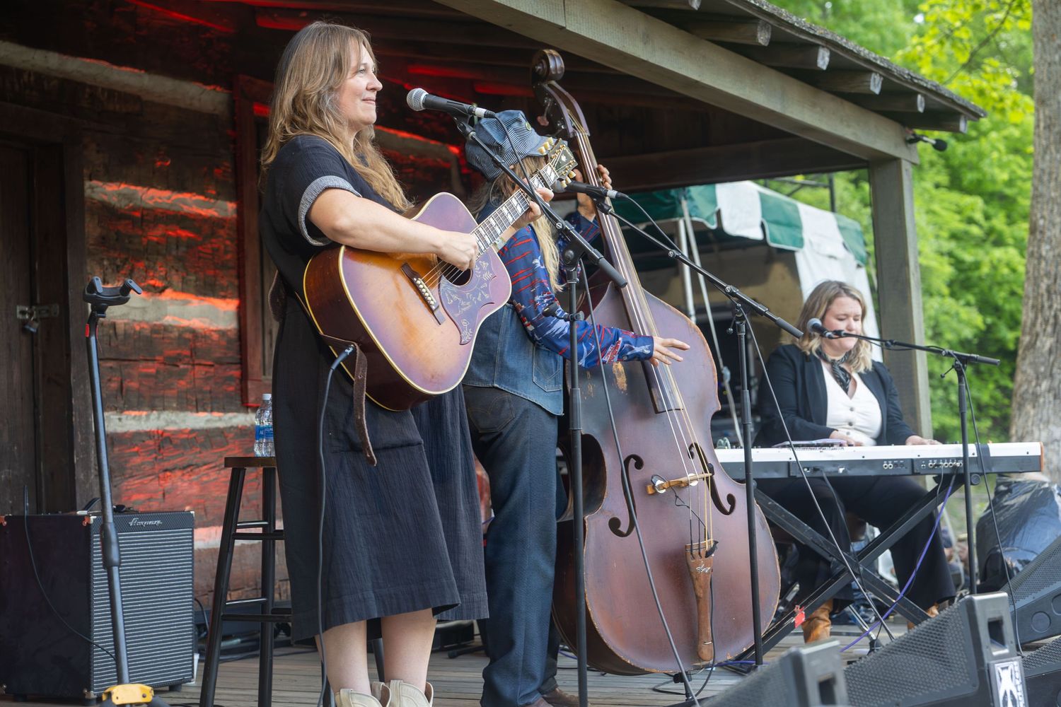 Live outdoor folk band performance with female guitarist at mic, upright bass player, and keyboardist on rustic stage.