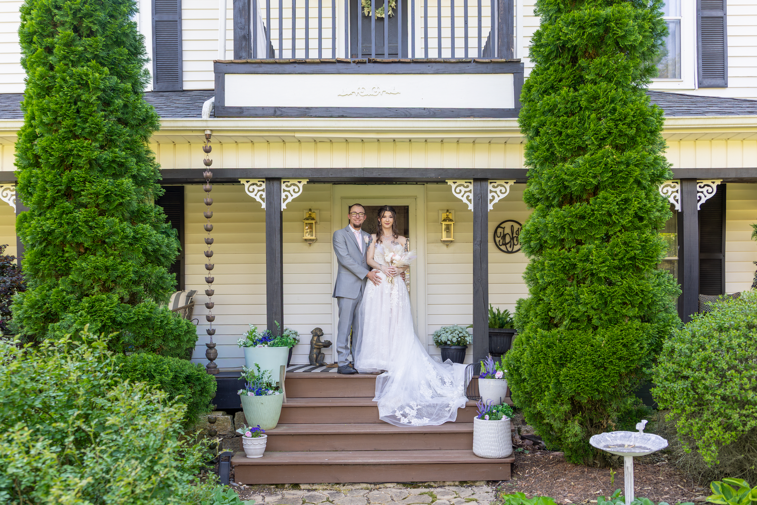 Bride and groom pose on porch steps of charming yellow house flanked by tall evergreen trees on wedding day.