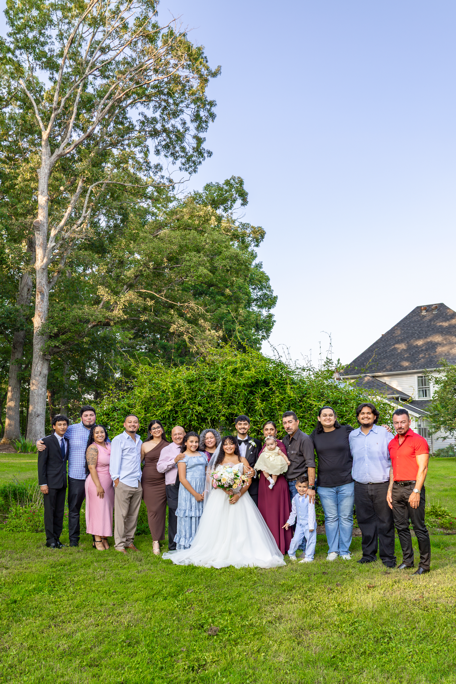 Wedding party poses outdoors on green lawn with tall trees and blue sky, bride in white gown holding bouquet.