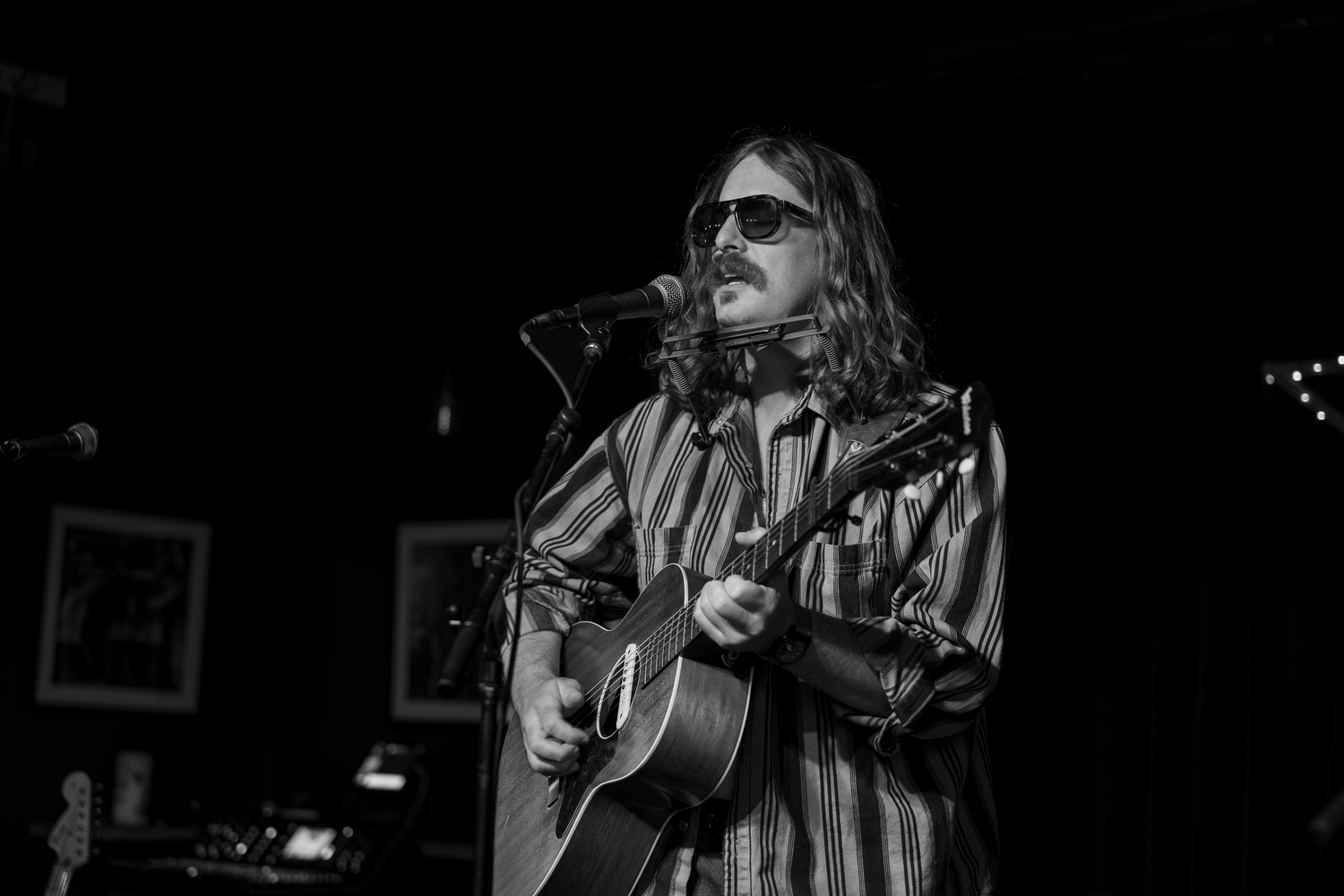 Musician with long hair and sunglasses playing acoustic guitar at a microphone on a dimly lit stage.
