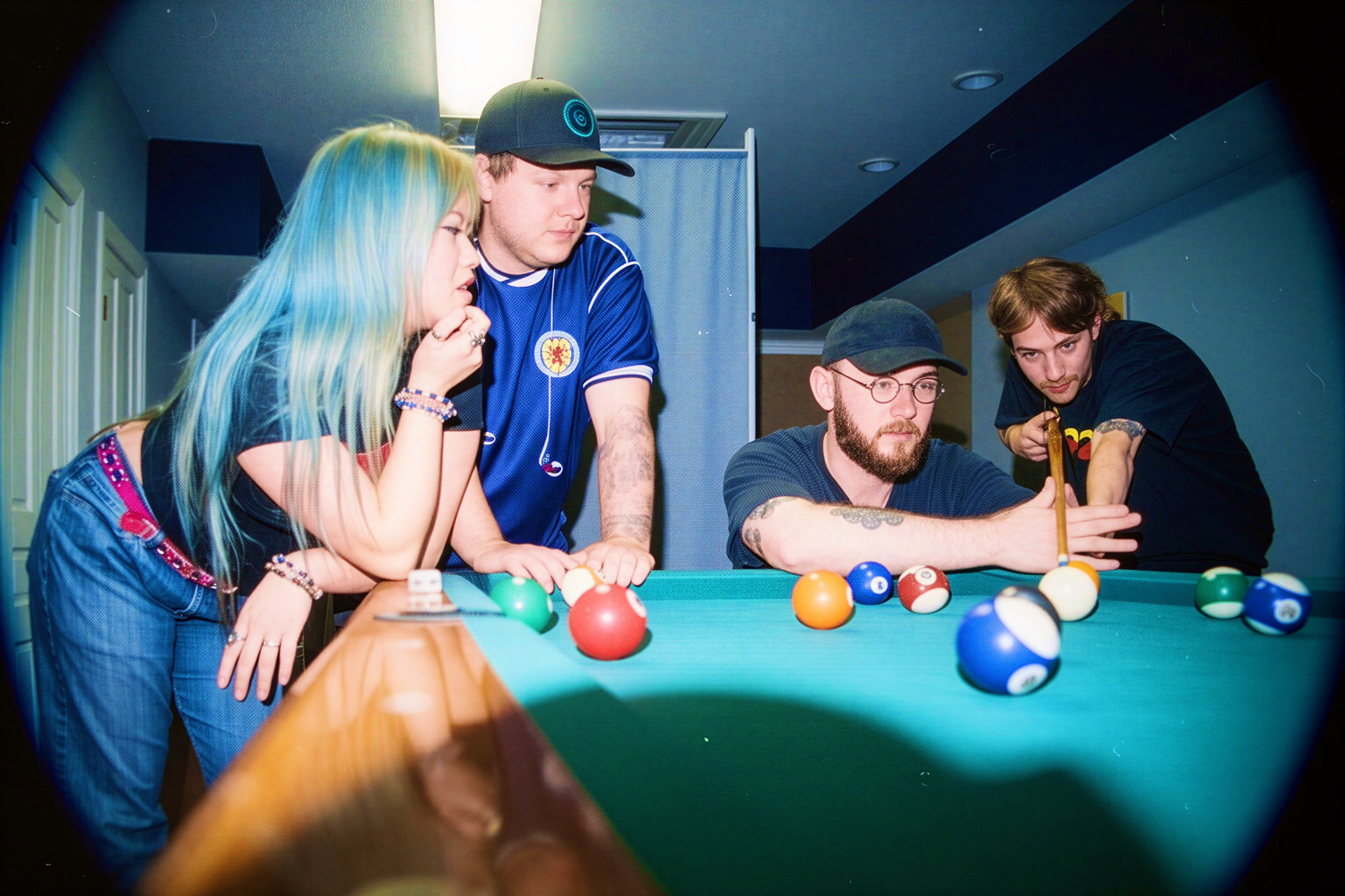 Four young adults playing pool in a game room, with colorful billiard balls on a green felt table.