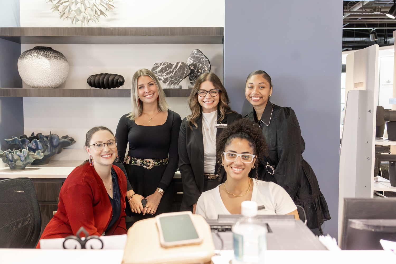 Five women smile together at a modern retail counter with decorative home decor items displayed on shelves behind them.