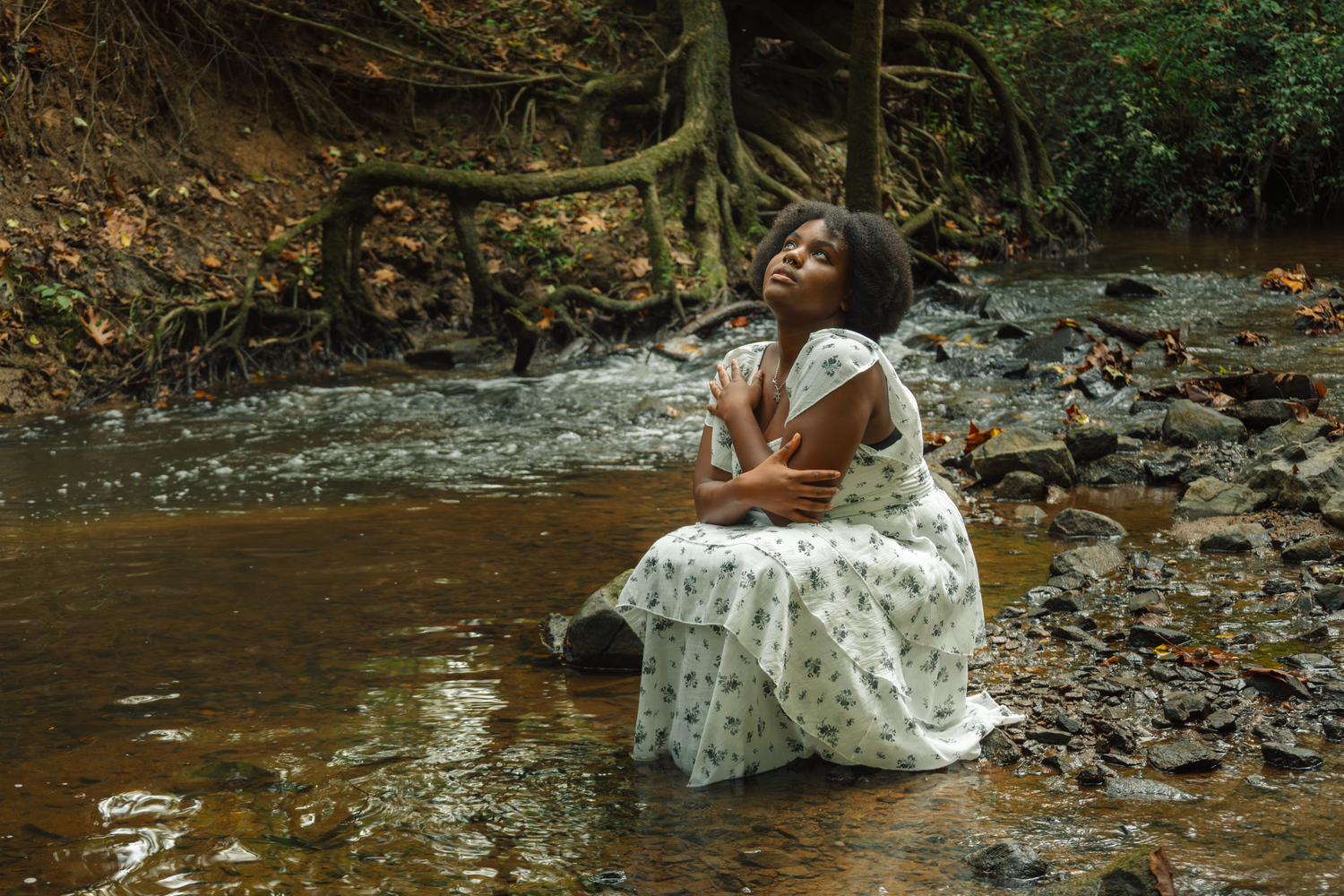 Woman in white dress sitting gracefully near a flowing stream in a forest setting.