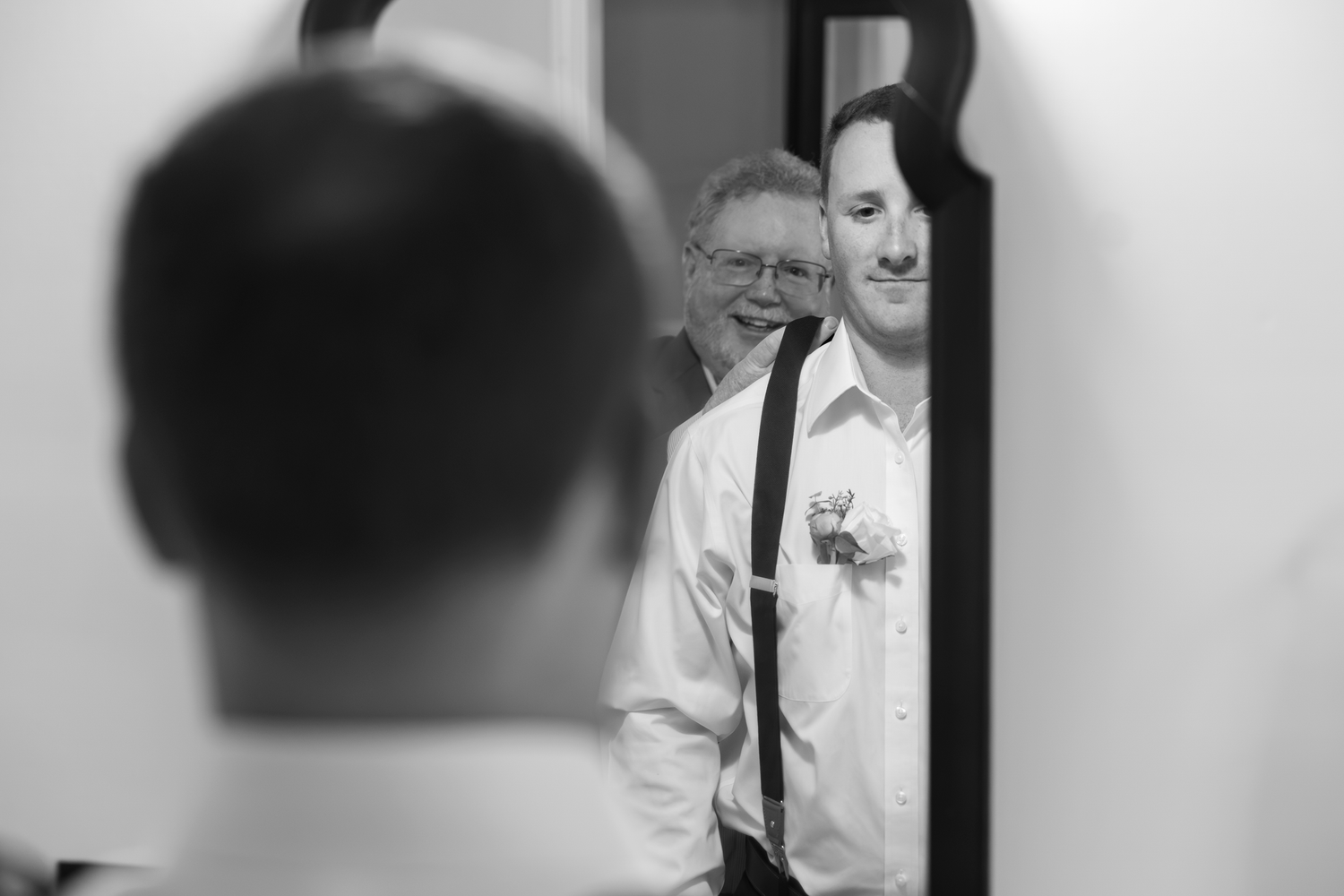 Black and white photo of groom and father smiling together in mirror reflection before wedding ceremony.
