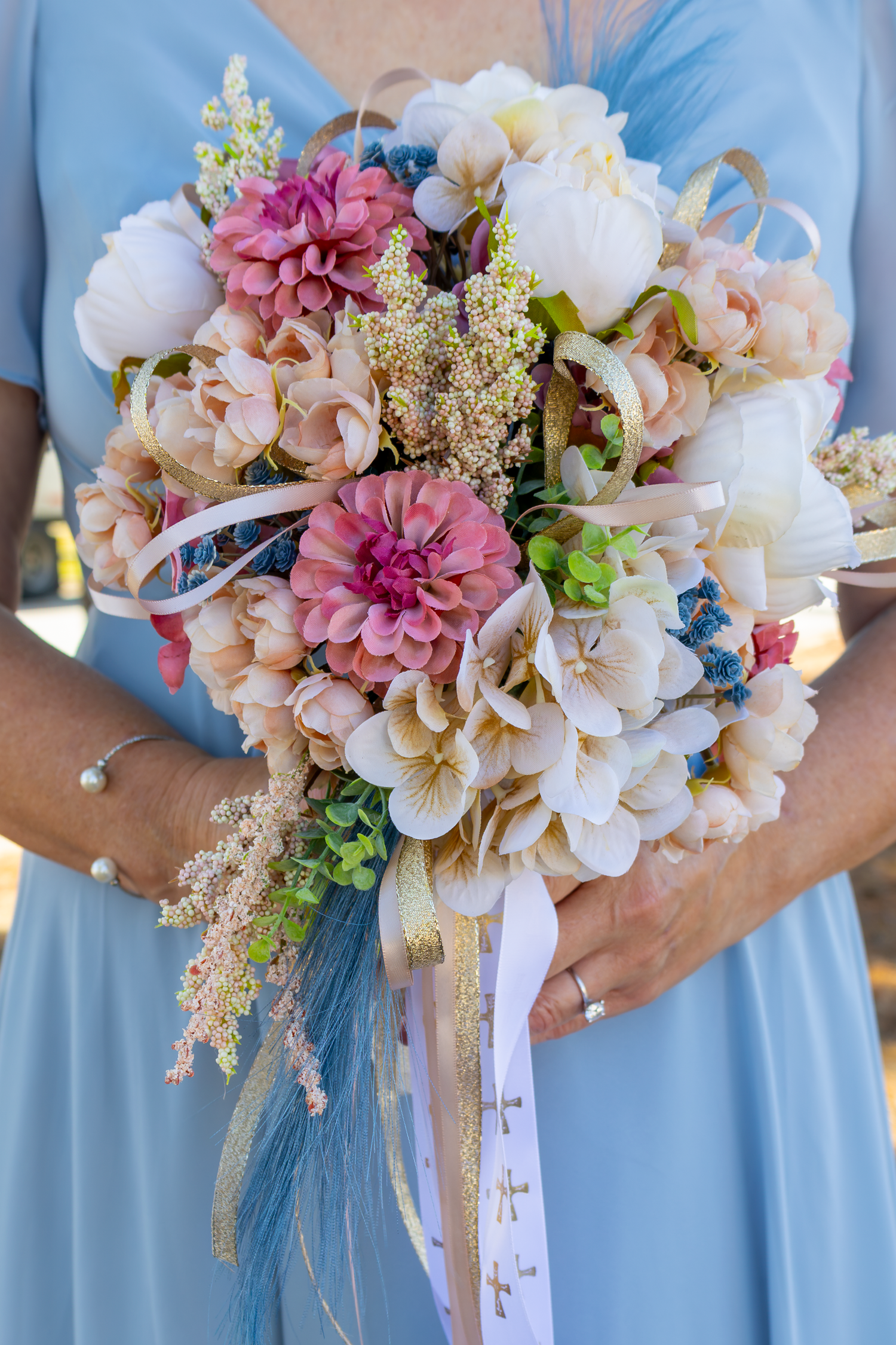 A bridesmaid in a light blue dress holds a colorful wedding bouquet with pink dahlias, white blooms, and ribbon accents.