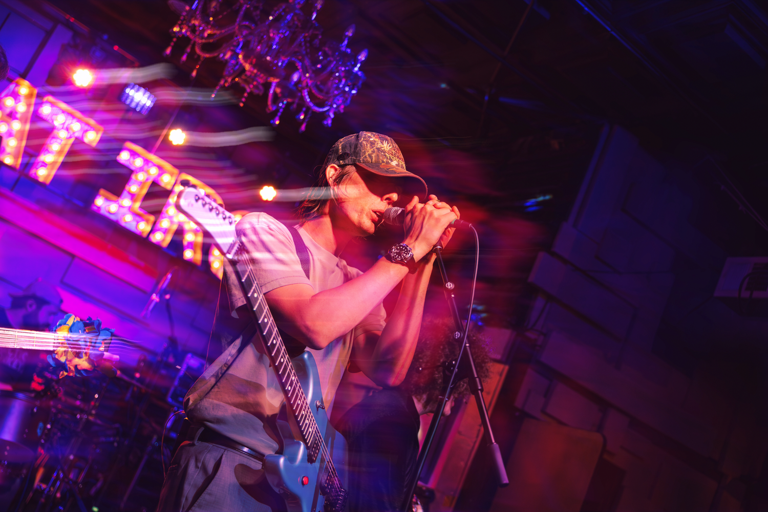Male guitarist singing into microphone on colorful purple and red lit stage during live rock concert performance.