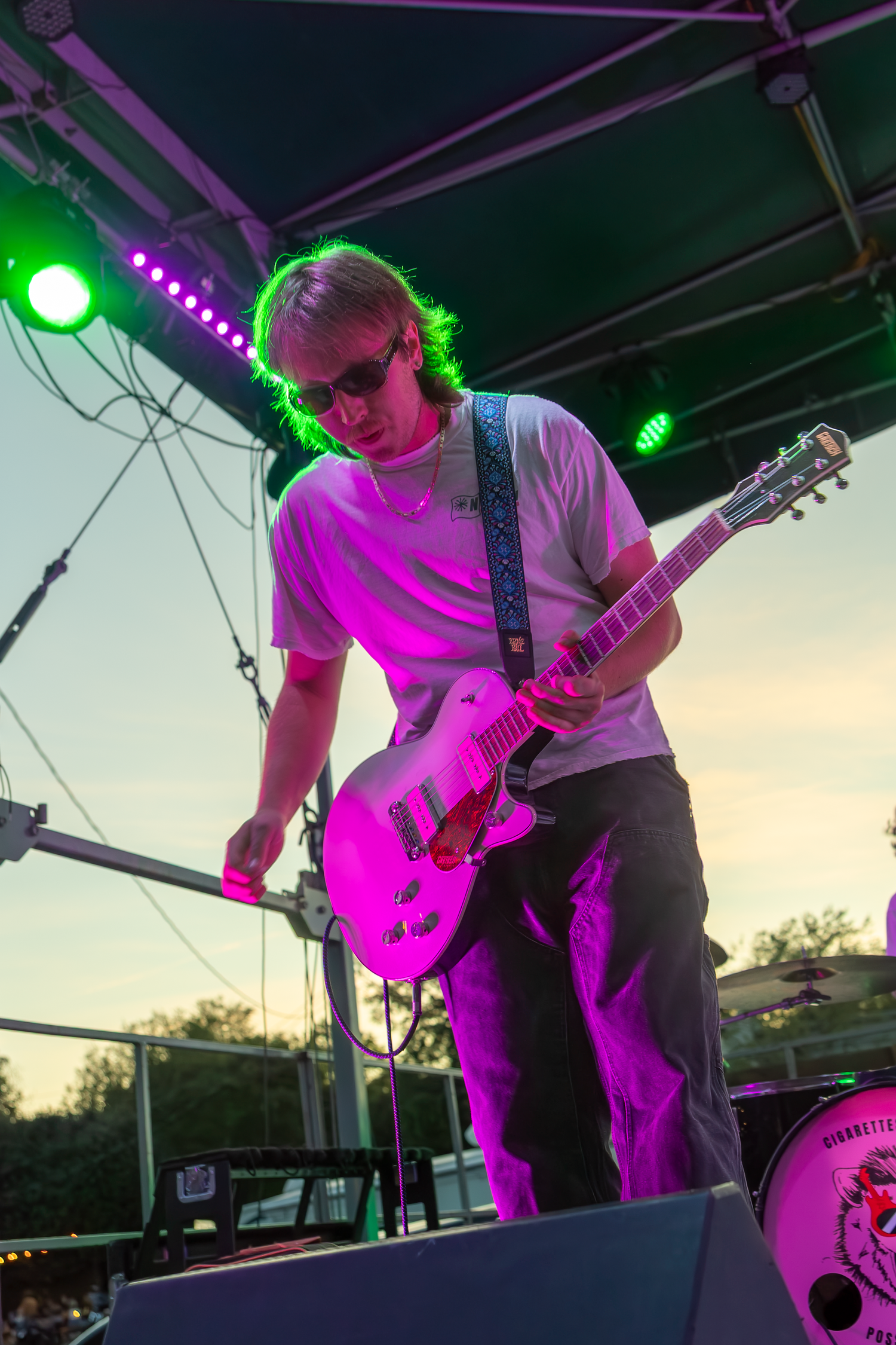 A guitarist performs on an outdoor stage under pink and green lights, wearing sunglasses and a white t-shirt at dusk.