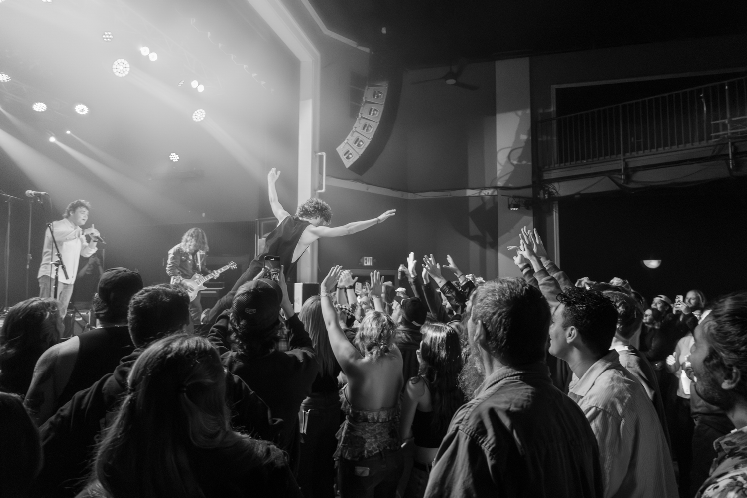 Black and white concert photo showing energetic crowd and performers on stage with dramatic lighting, as one performer leans to crowdsurf.