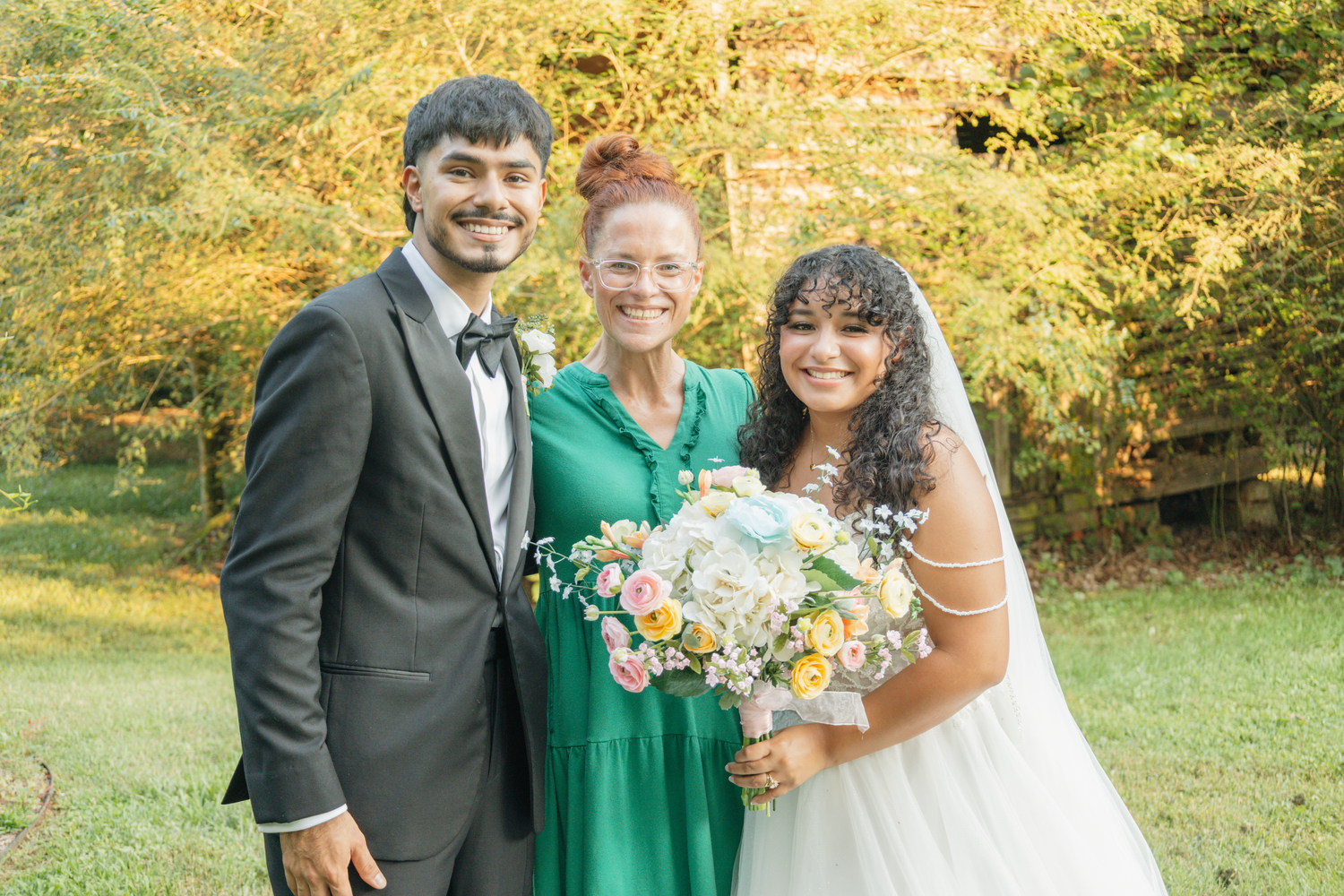 A joyful wedding photo featuring a couple and guest in formal attire with autumn foliage backdrop at The Farmhouse Siloam.
