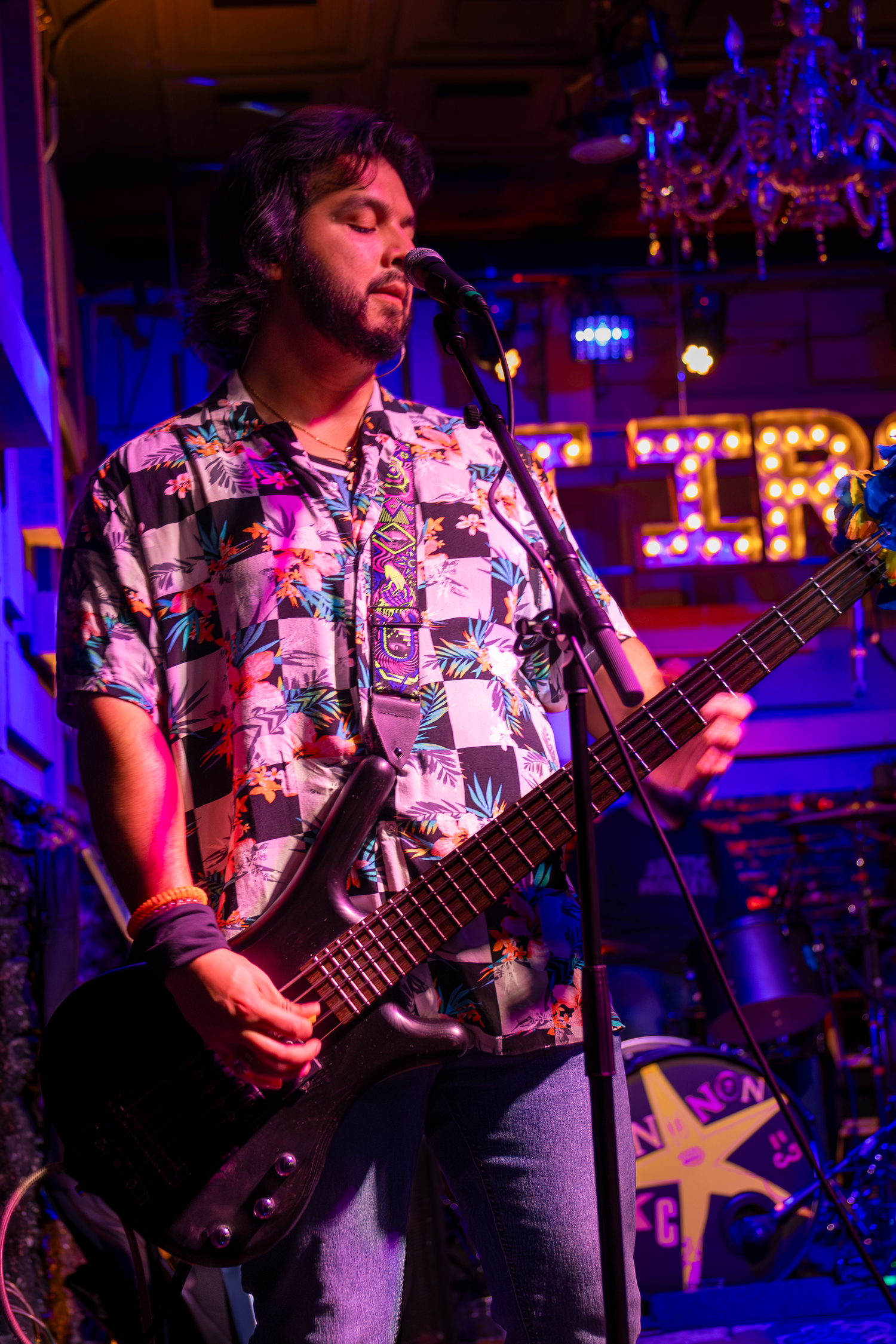 A guitarist in a colorful Hawaiian shirt performs on stage under purple lights at a lively music venue.