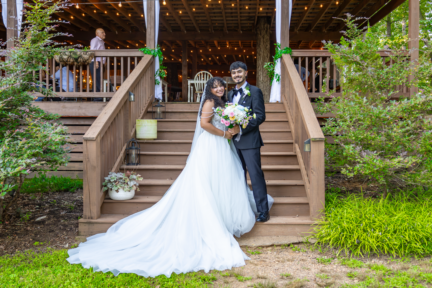 Bride and groom pose on wooden staircase at outdoor wedding venue with string lights and lush greenery.