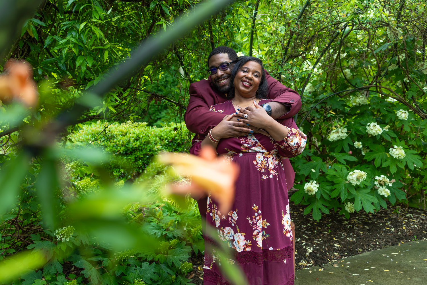 A couple in matching burgundy outfits embrace joyfully in a lush green garden with white flowering bushes.