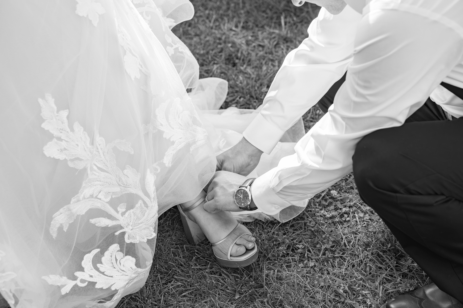 Black and white photo of groom kneeling to fasten bride's wedding shoe on grass outdoor ceremony.