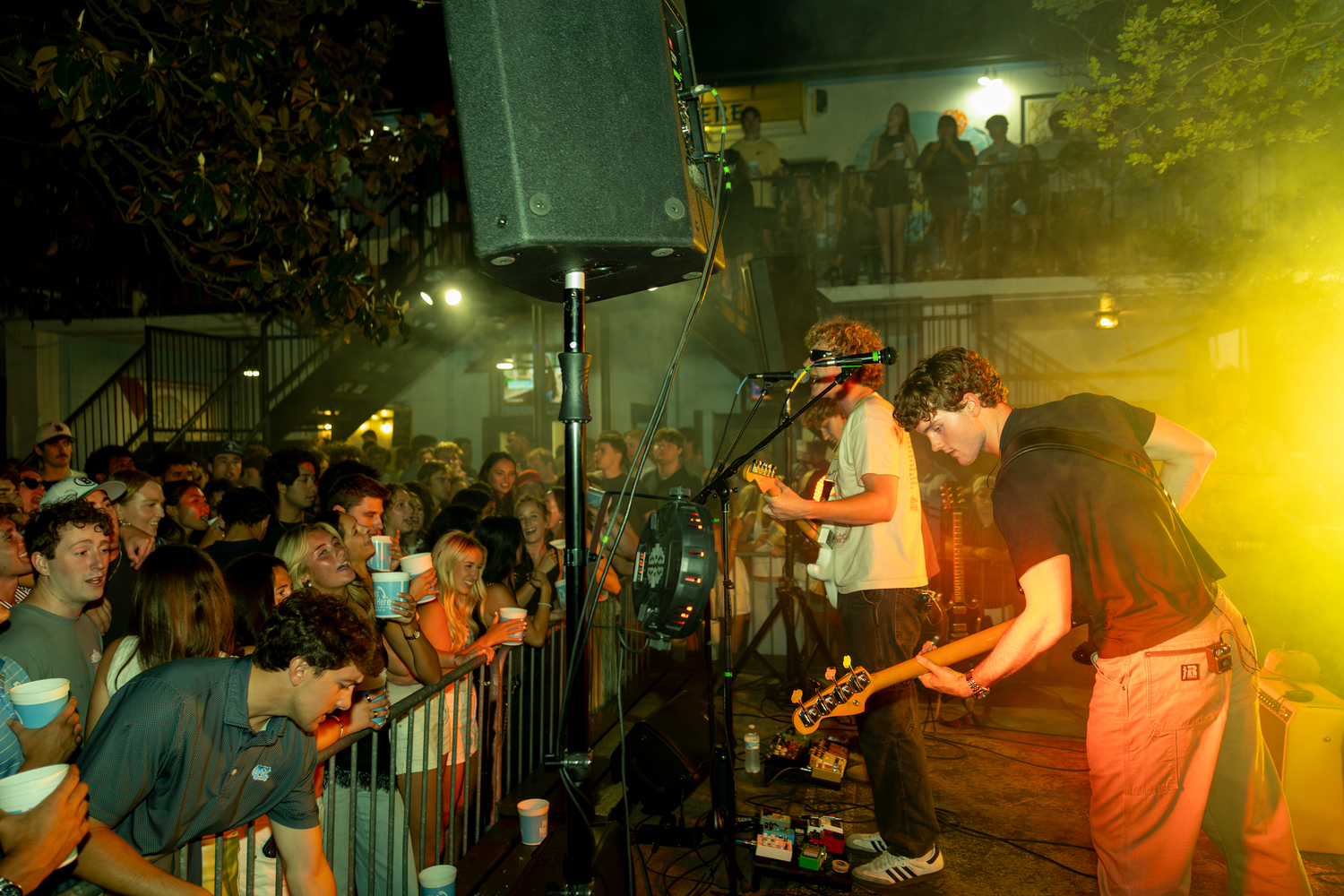 Live band performing on outdoor stage at night with large crowd gathered behind metal barriers.