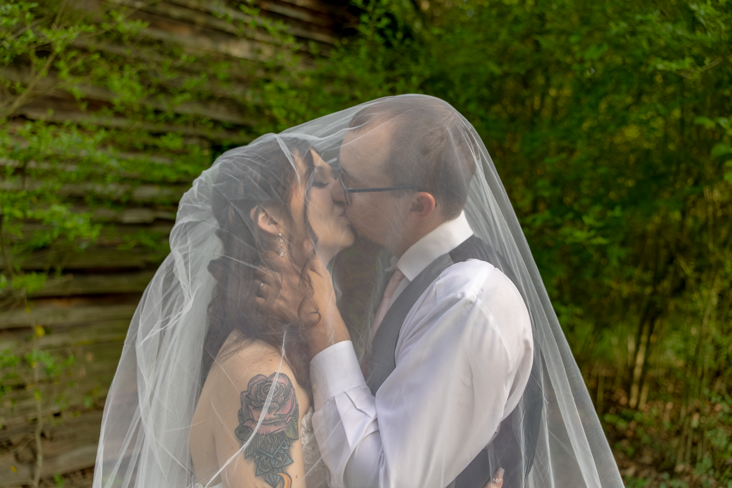 Bride and groom sharing a romantic kiss under a wedding veil outdoors surrounded by lush green trees.