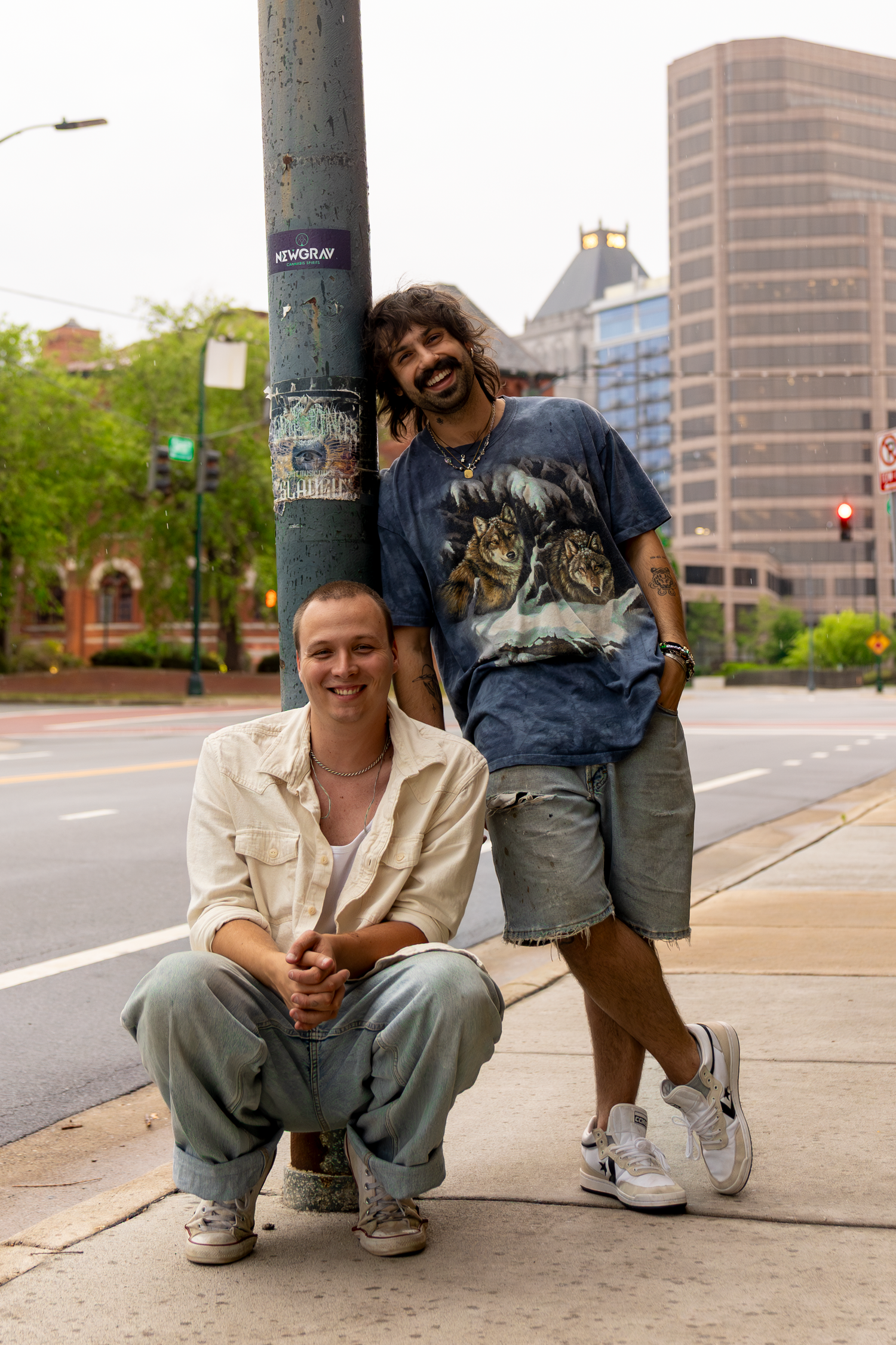 Two men pose on a city sidewalk near a light pole, one crouching and one standing, in casual streetwear.
