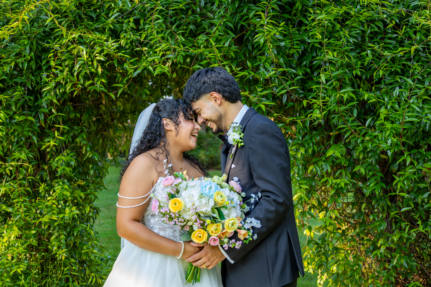 Bride and groom touching foreheads, smiling, holding colorful bouquet before lush green ivy wall.