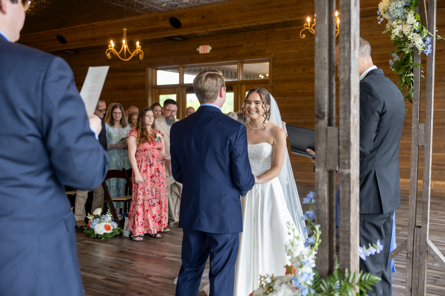 Bride smiling at groom during indoor rustic barn wedding ceremony with floral decorations and guests in background.
