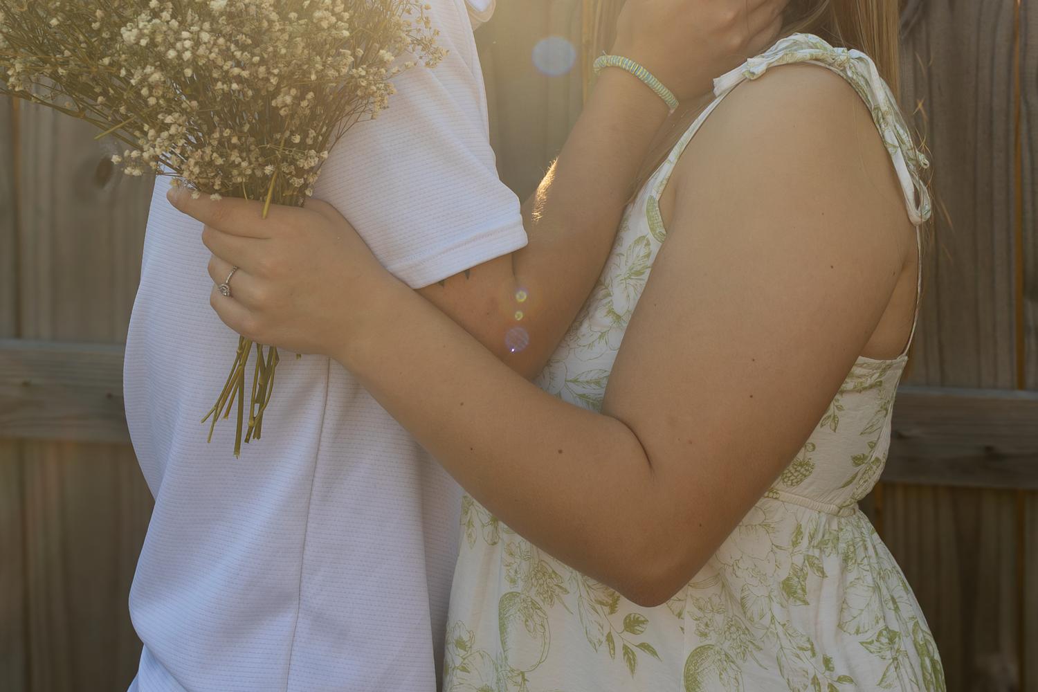 A couple holds a delicate bouquet of baby's breath flowers against a wooden fence in soft sunlight.
