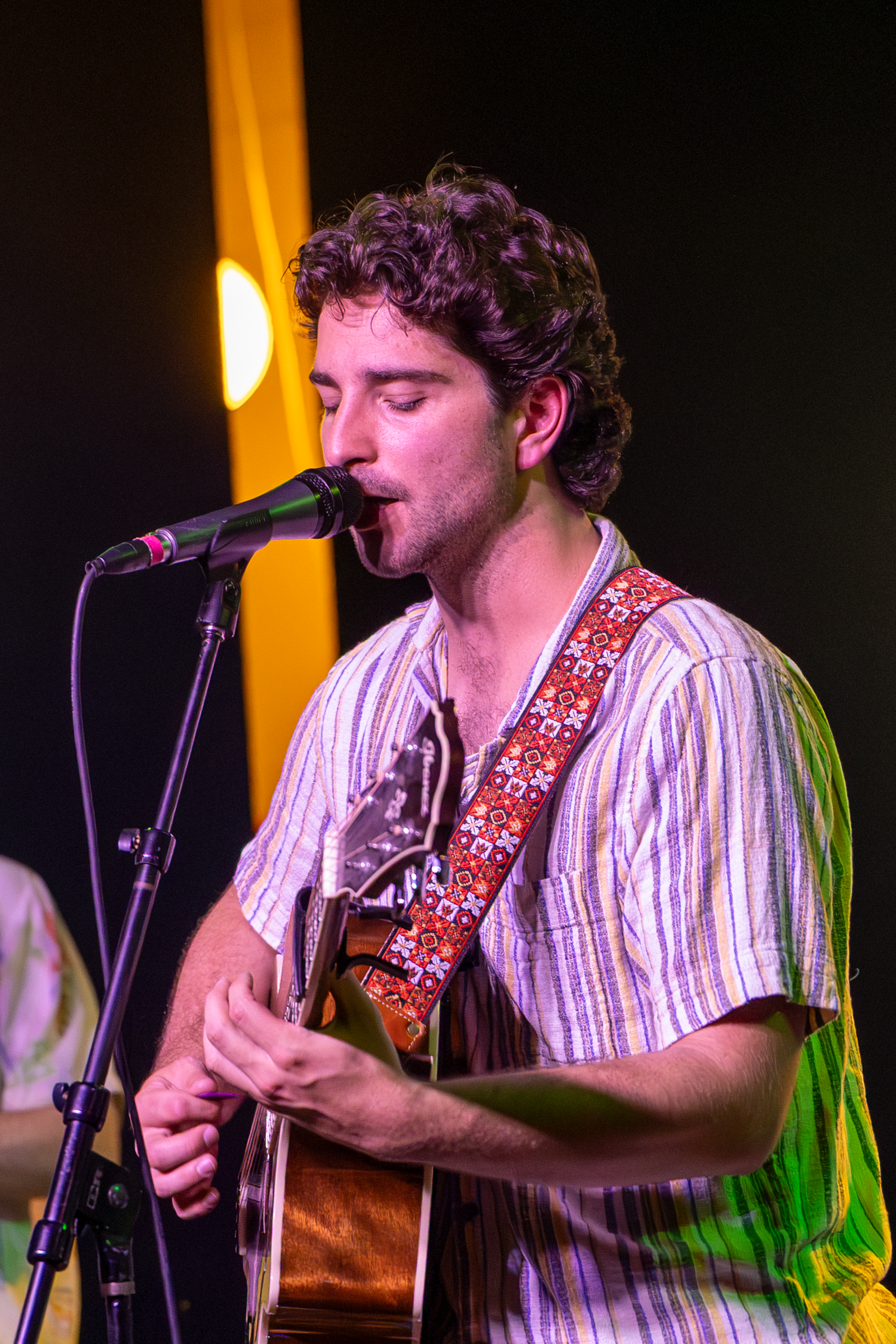 Musician performs on stage with acoustic guitar and microphone wearing striped shirt and decorative guitar strap.