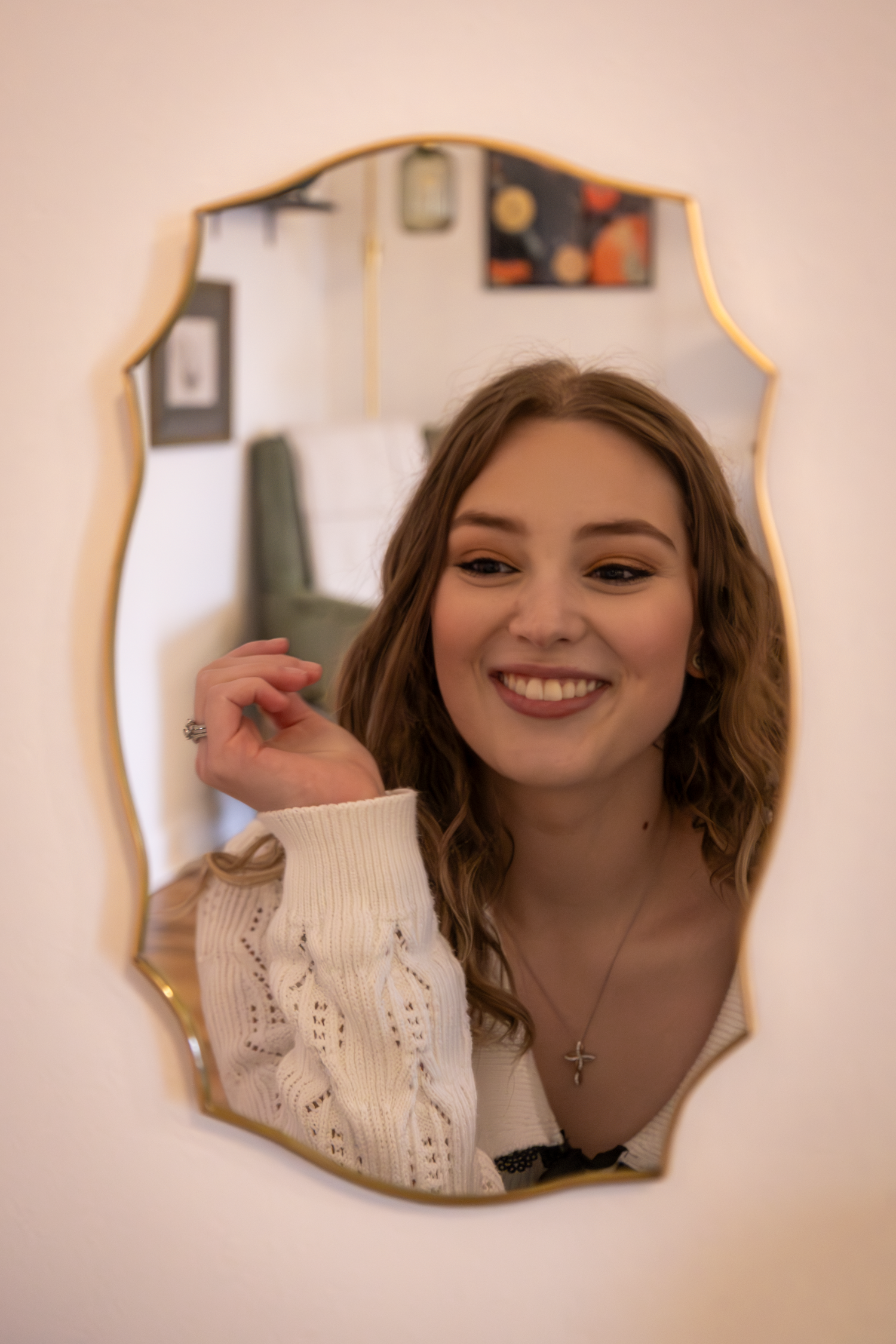 A reflection in an ornate gold mirror shows someone in a white sweater smiling.