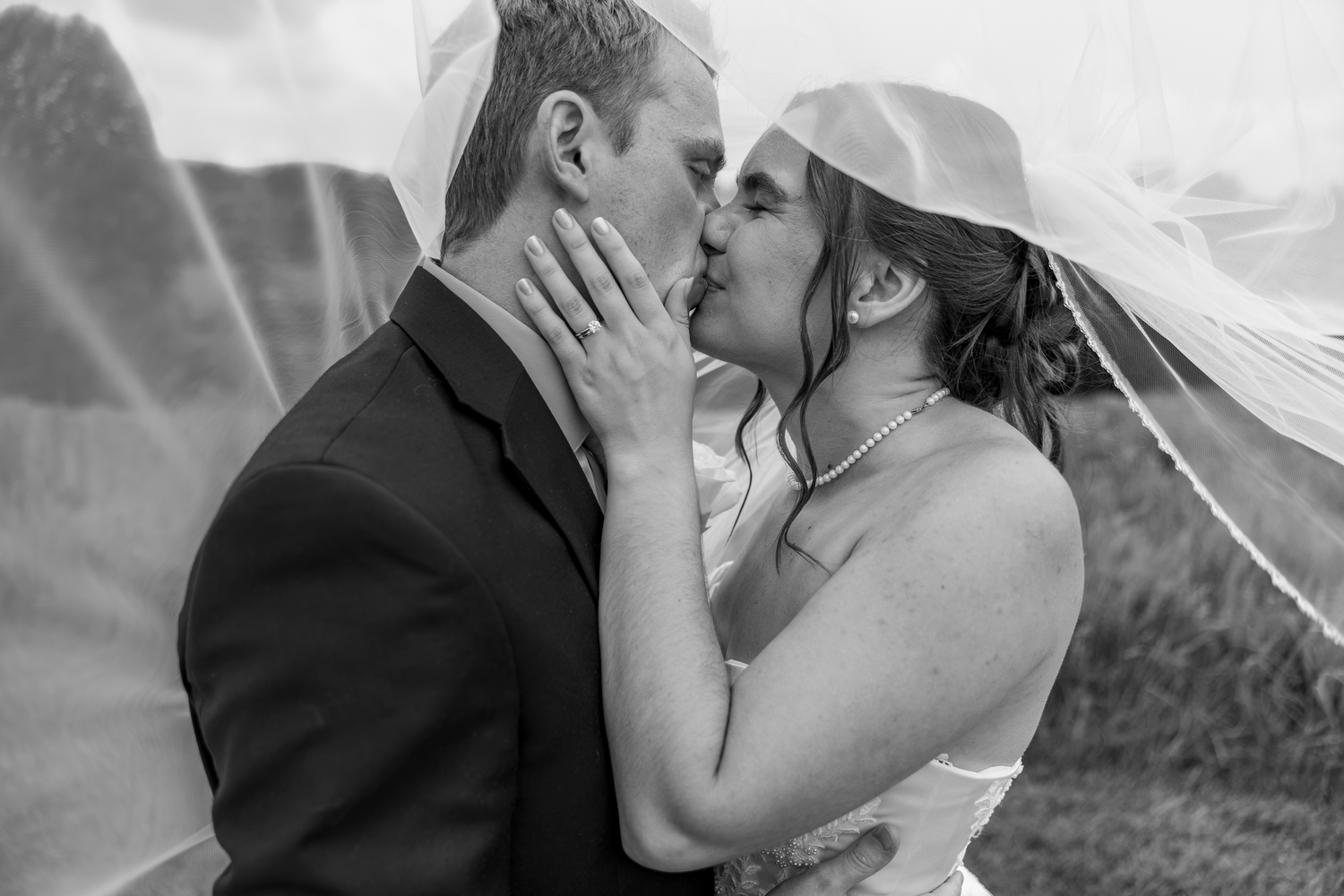 Black and white photo of a bride and groom sharing a romantic kiss under a flowing veil on their wedding day.