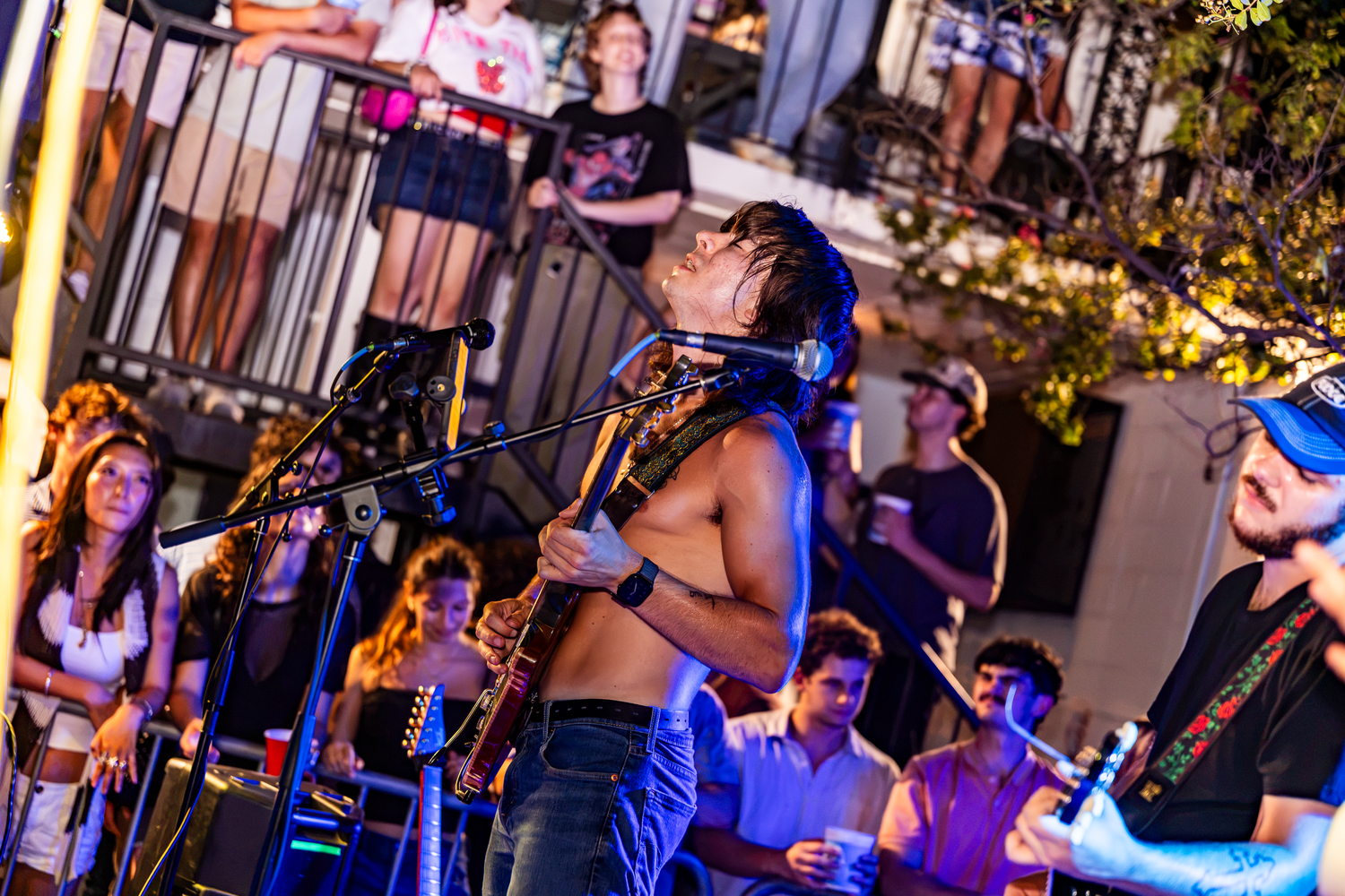 Shirtless guitarist performs outdoors at a lively night concert with an engaged crowd watching nearby.
