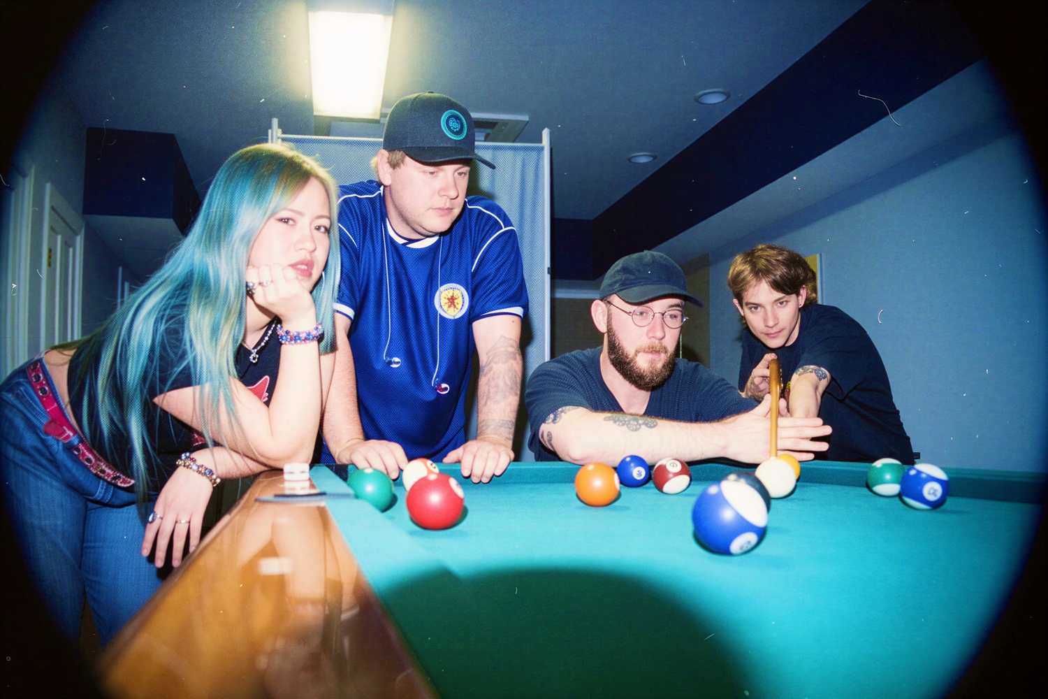Four young adults posing around a pool table with colorful billiard balls in a game room.
