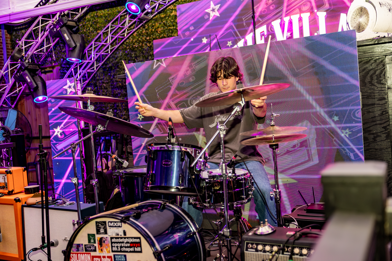 Drummer performing energetically on stage with colorful laser lights and a vibrant LED backdrop at a live music venue.