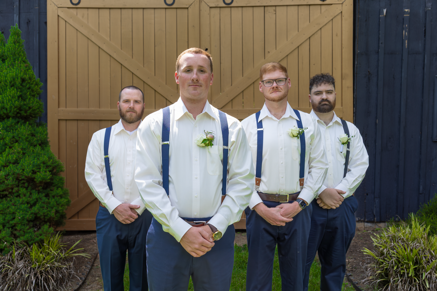 Four groomsmen in white shirts and blue suspenders pose in front of rustic barn doors at a wedding.