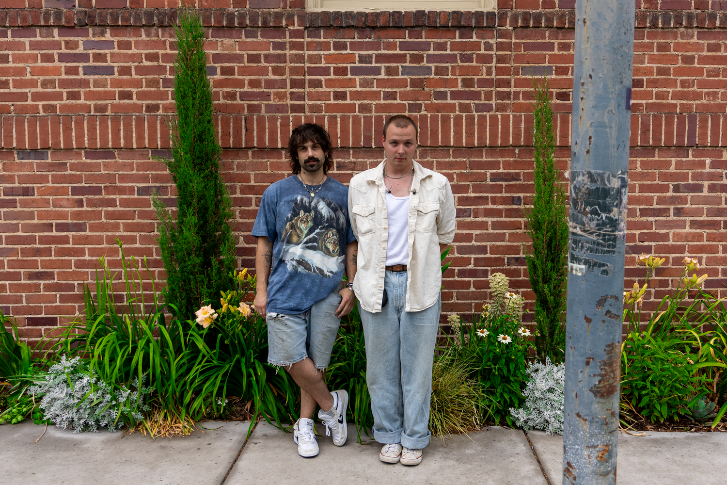 Two men standing in front of a brick wall with cypress trees and flowers in an outdoor setting.