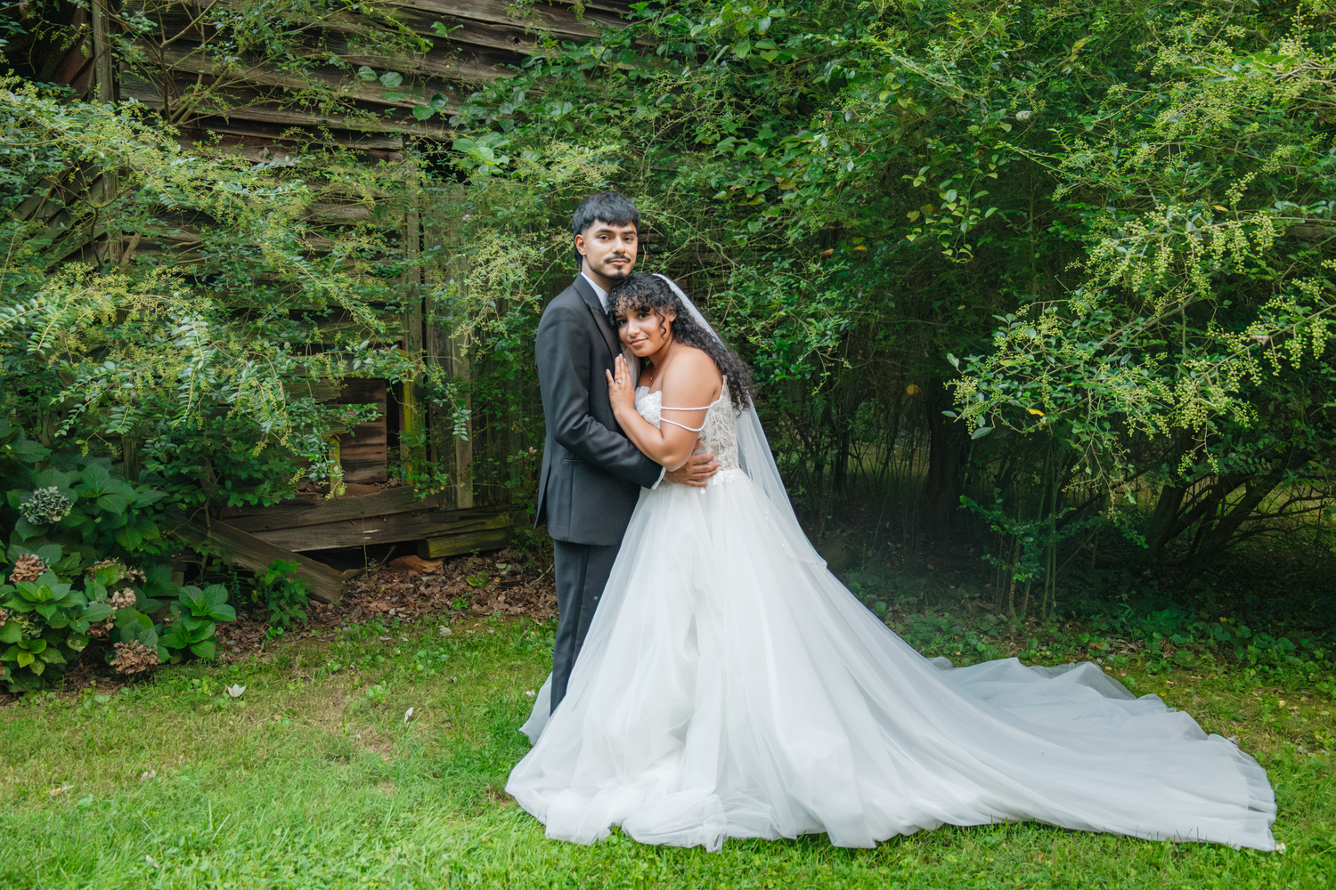 Bride in white gown with long train embraces groom in gray suit outdoors near rustic wooden structure surrounded by lush greenery.