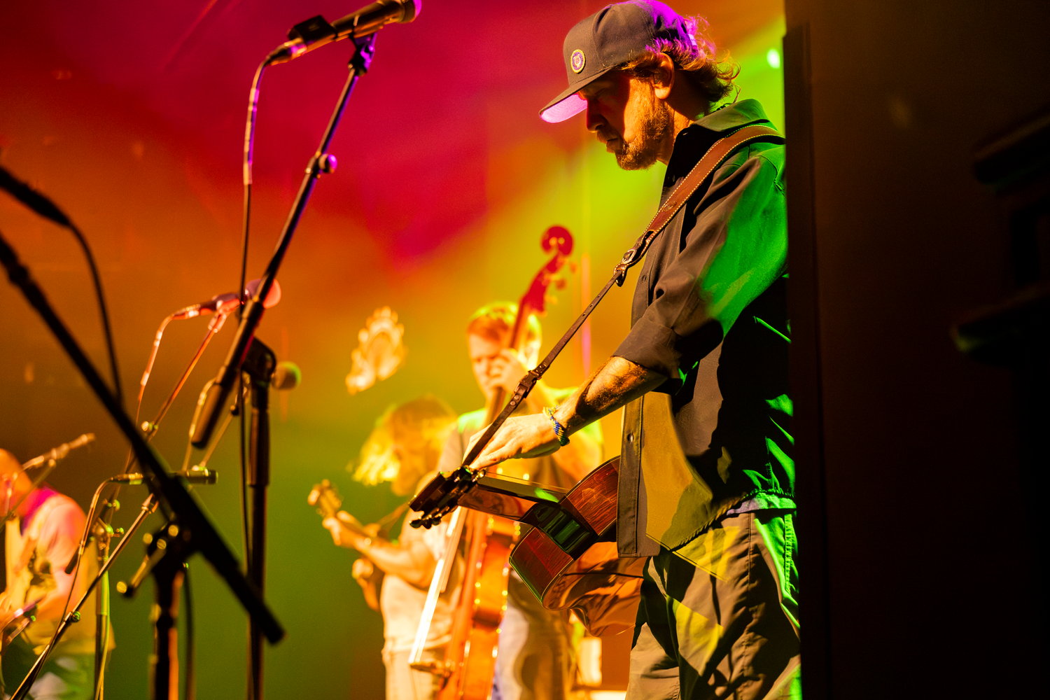 Live band performs on colorful stage with guitarist in cap at microphone under red and green lights.