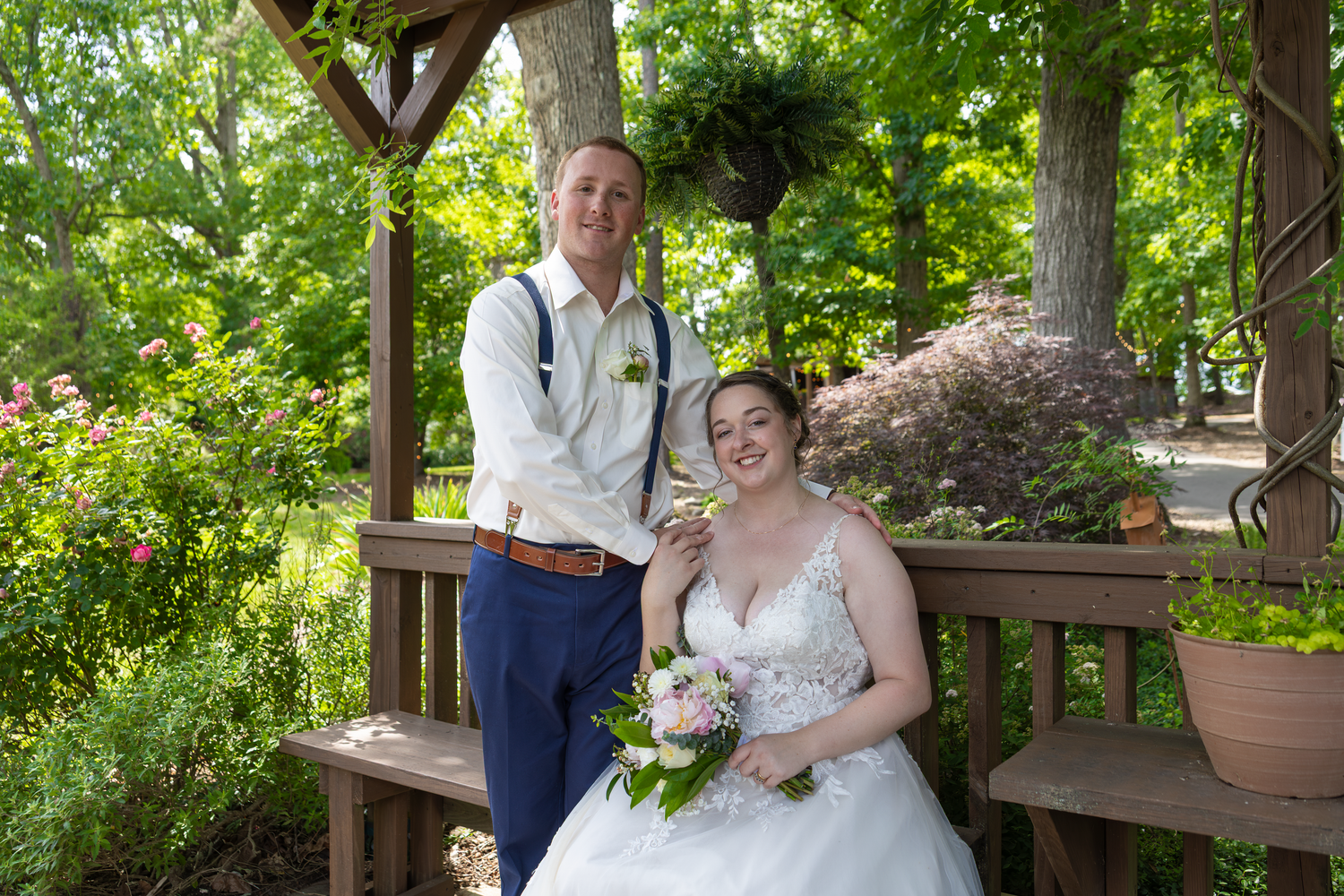 A bride and groom pose together in a lush garden gazebo, bride holding a bouquet in a white gown.