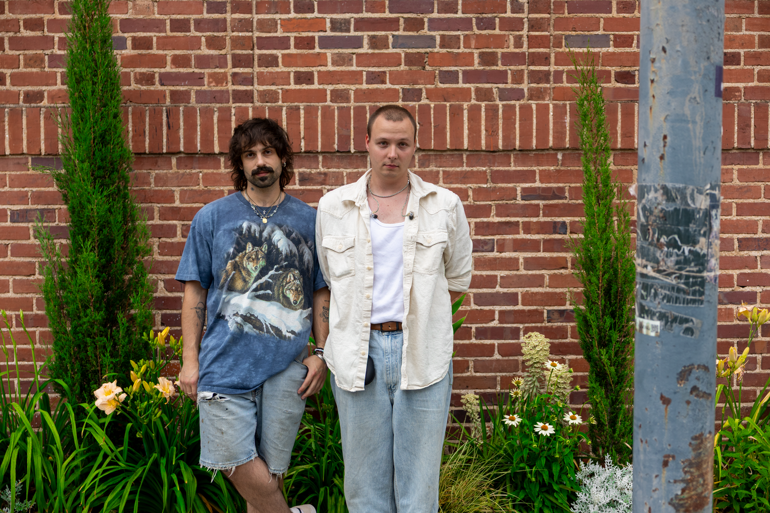 Two men standing against a brick wall with green shrubs and yellow flowers in an outdoor garden setting.