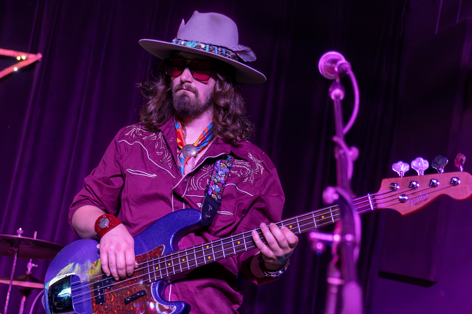 Musician in gray hat and purple western shirt plays bass guitar on a dimly lit stage with purple lighting.