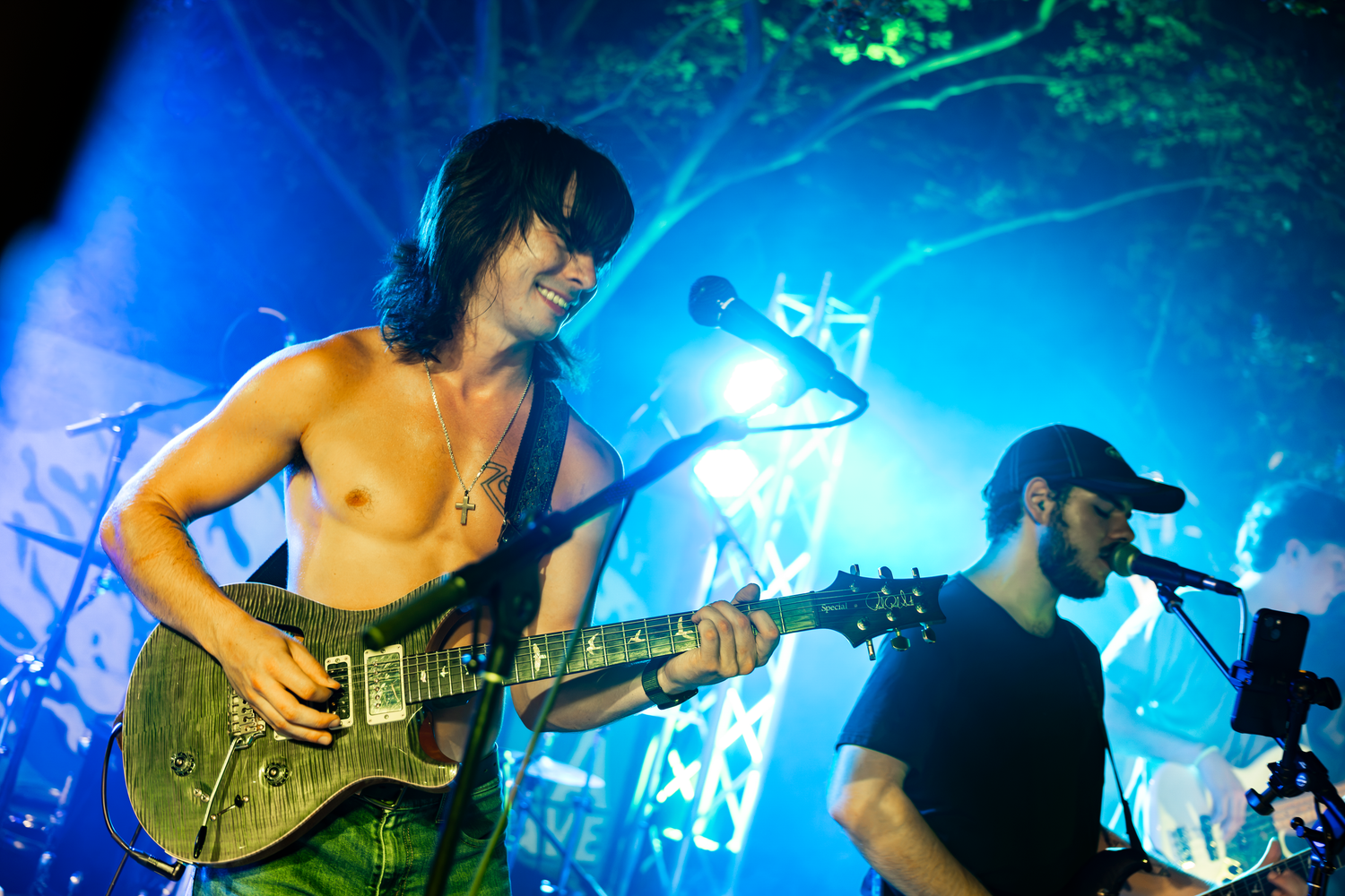 Shirtless guitarist performs on stage with blue lights at an outdoor concert alongside a bearded musician.